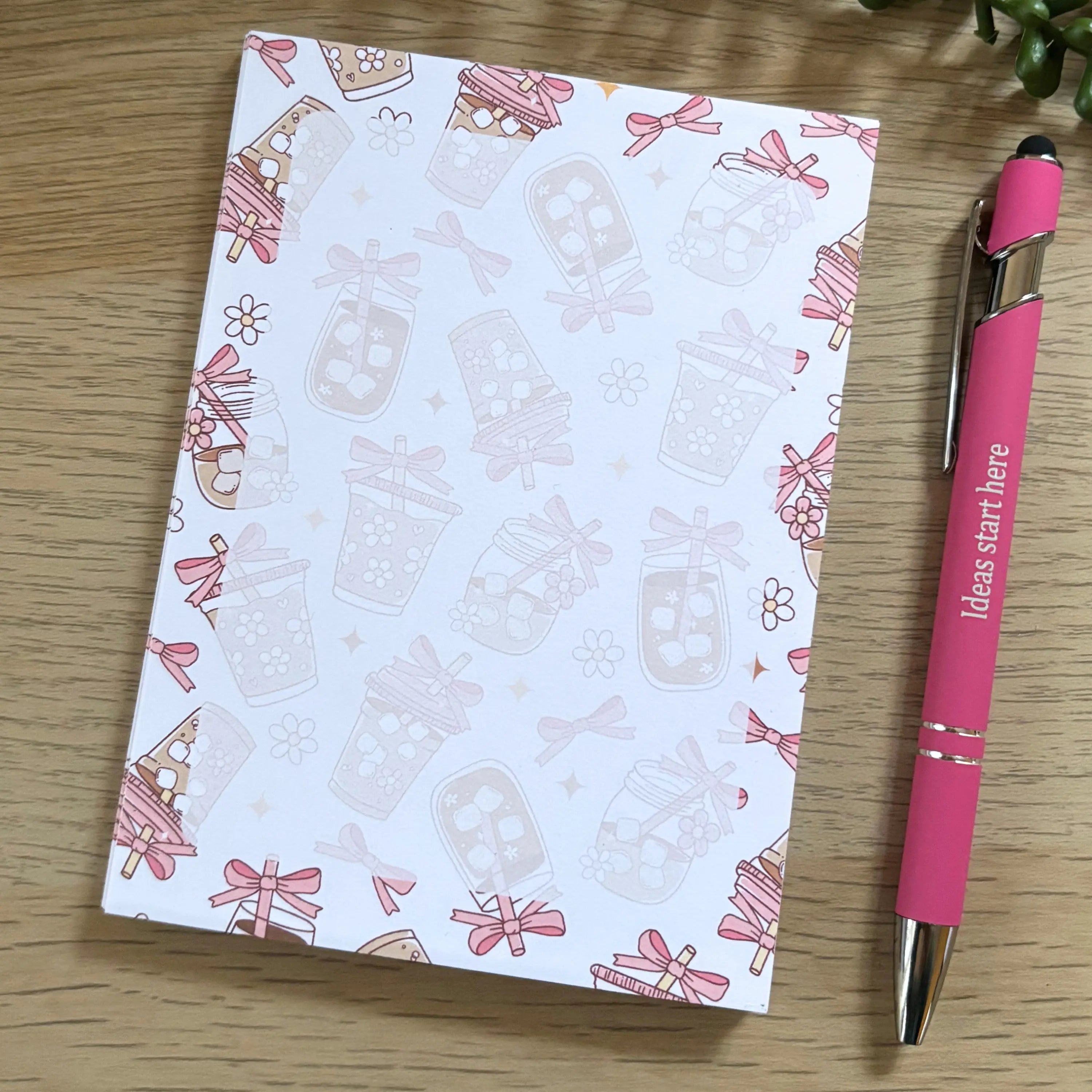 Notepad with pink iced coffee cup illustrations and small bows on a white background, placed on a wooden surface with a pink .