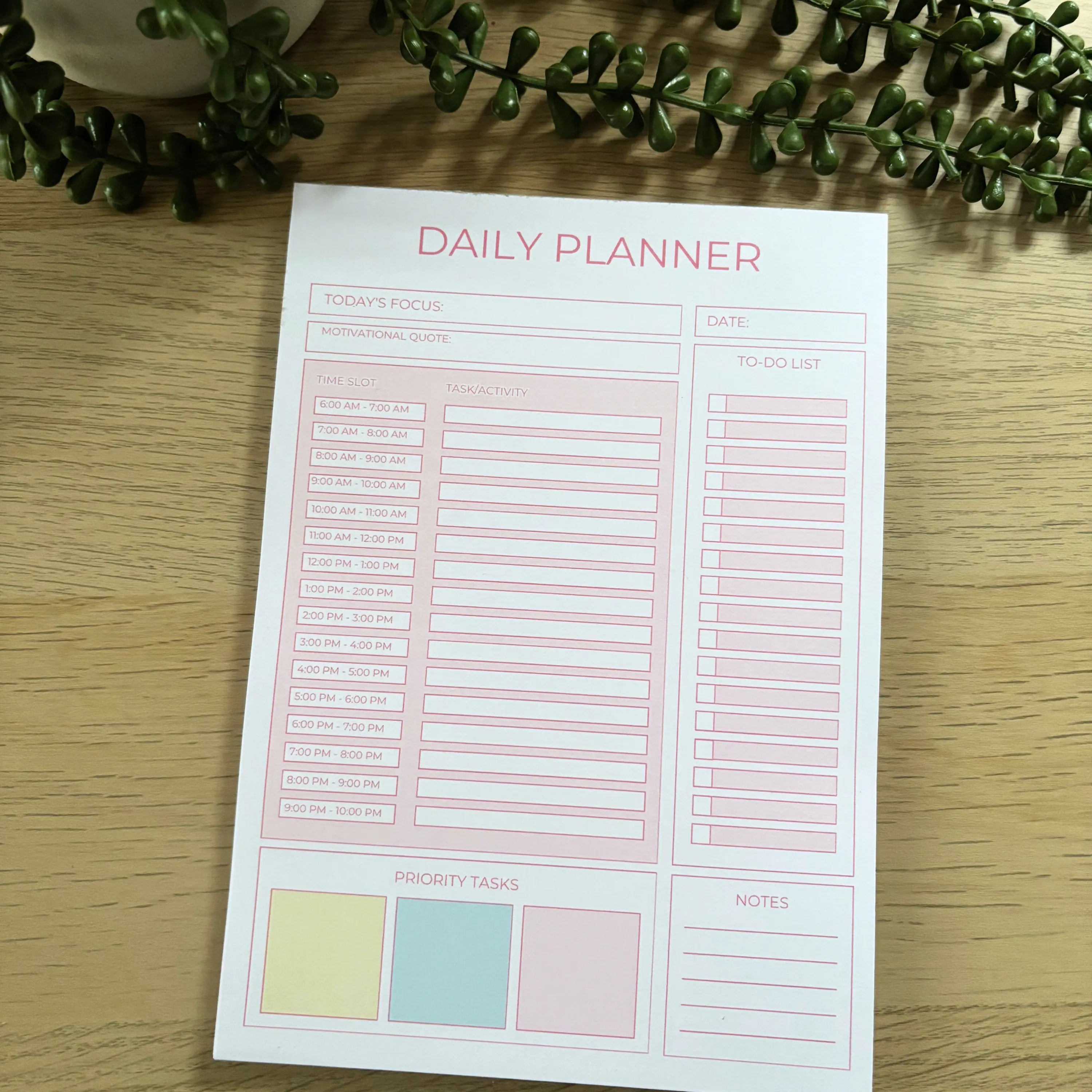 Minimalist pink daily planner notepad with time slots, to-do list, priority tasks, and notes sections on wooden surface.