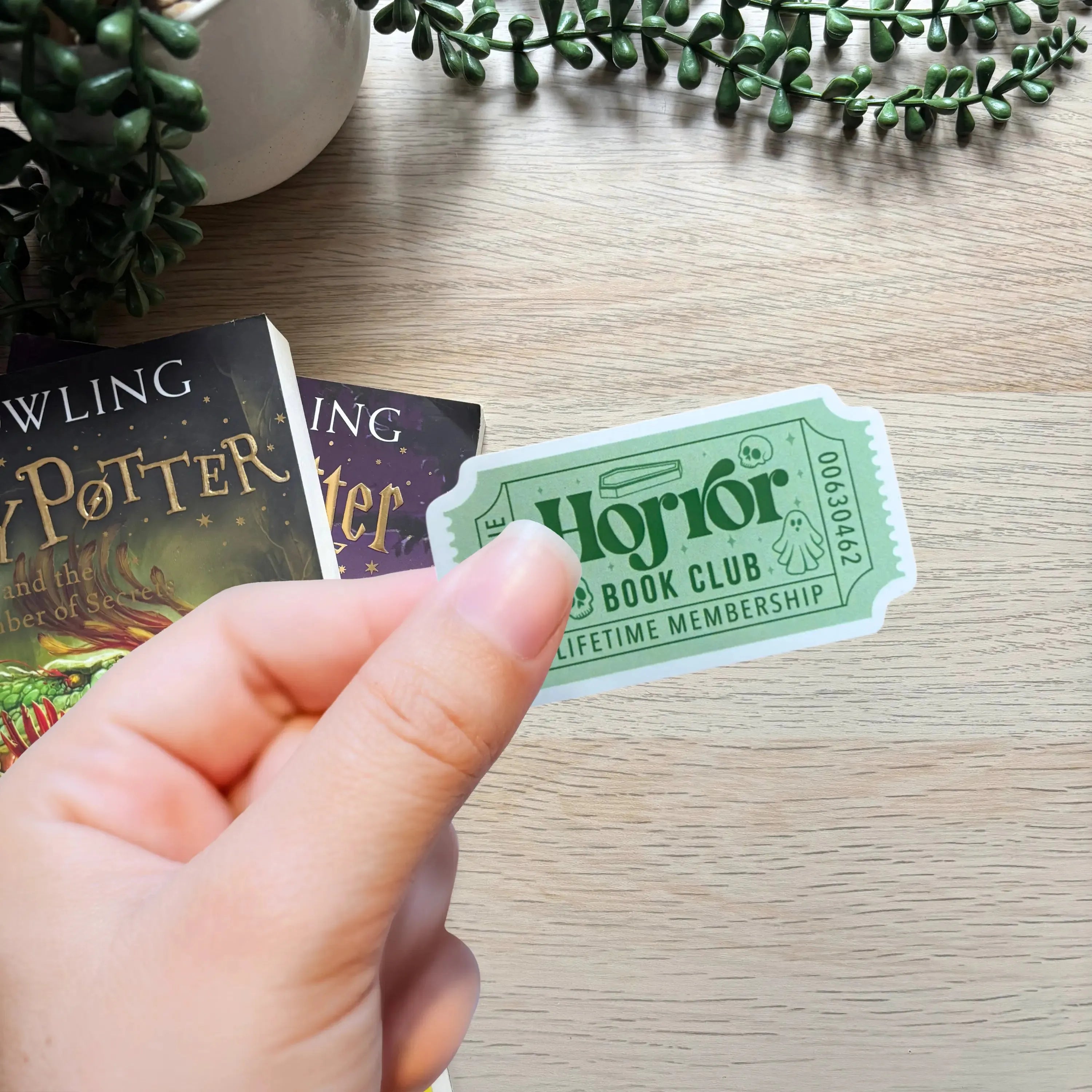 Green and black minimalist ticket-shaped sticker reading Horror Book Club Lifetime Membership held by hand over wooden.