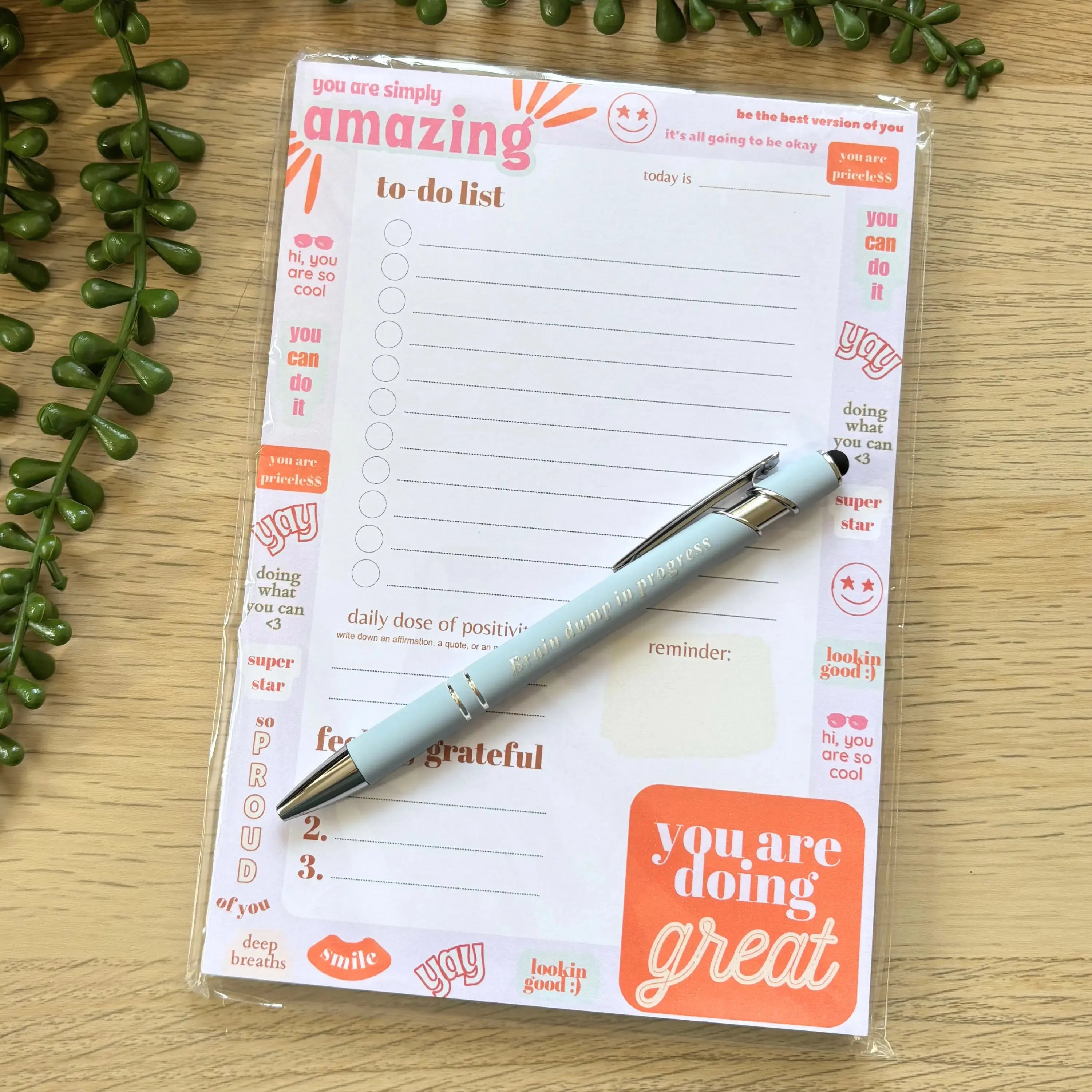 Minimalist to-do list notepad with "You are amazing" header and motivational orange accents with pen on wooden surface.
