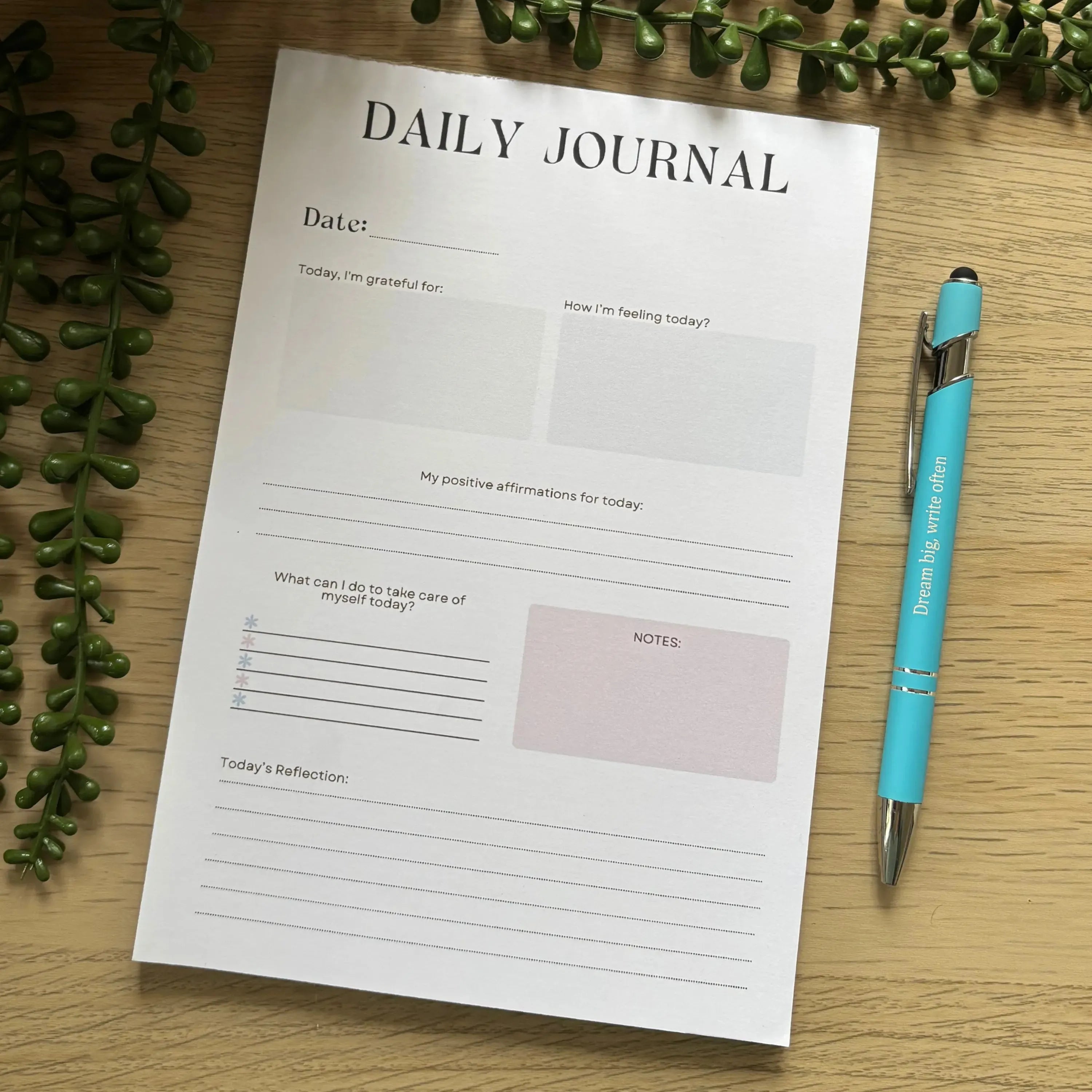 Minimalist daily journal notepad with sections for date, gratitude, goals, and reflections, shown with blue pen.