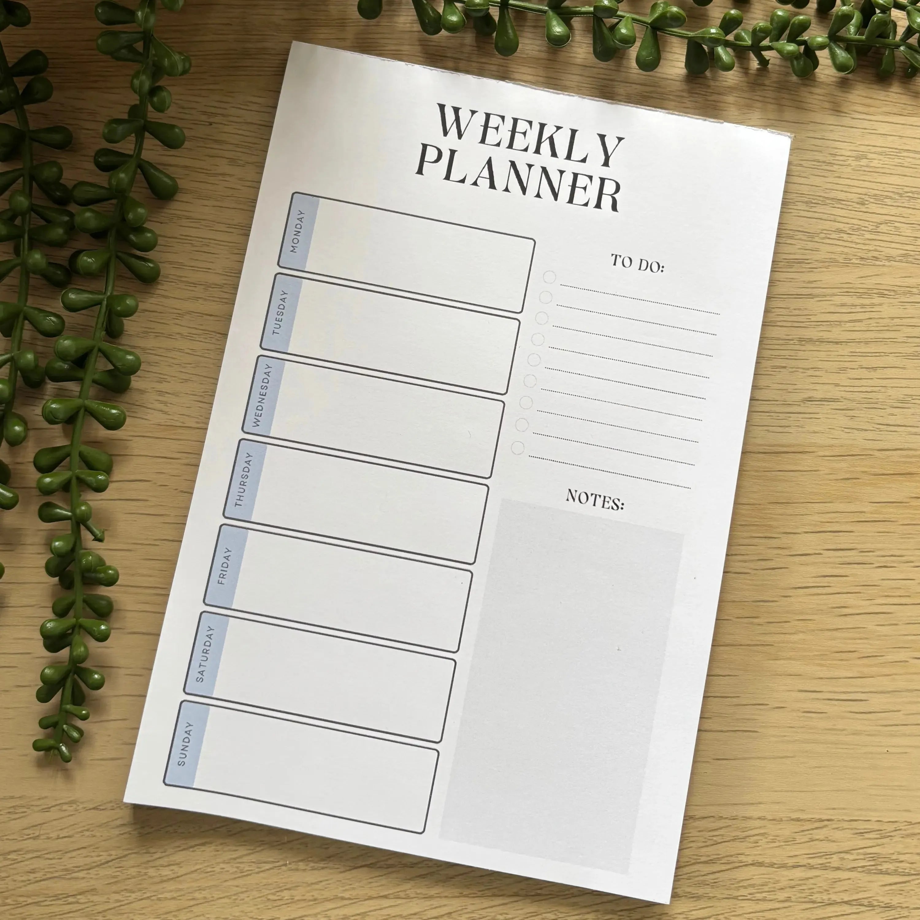Minimalist weekly planner notepad with blue day headers, to-do list, and notes section on white paper.