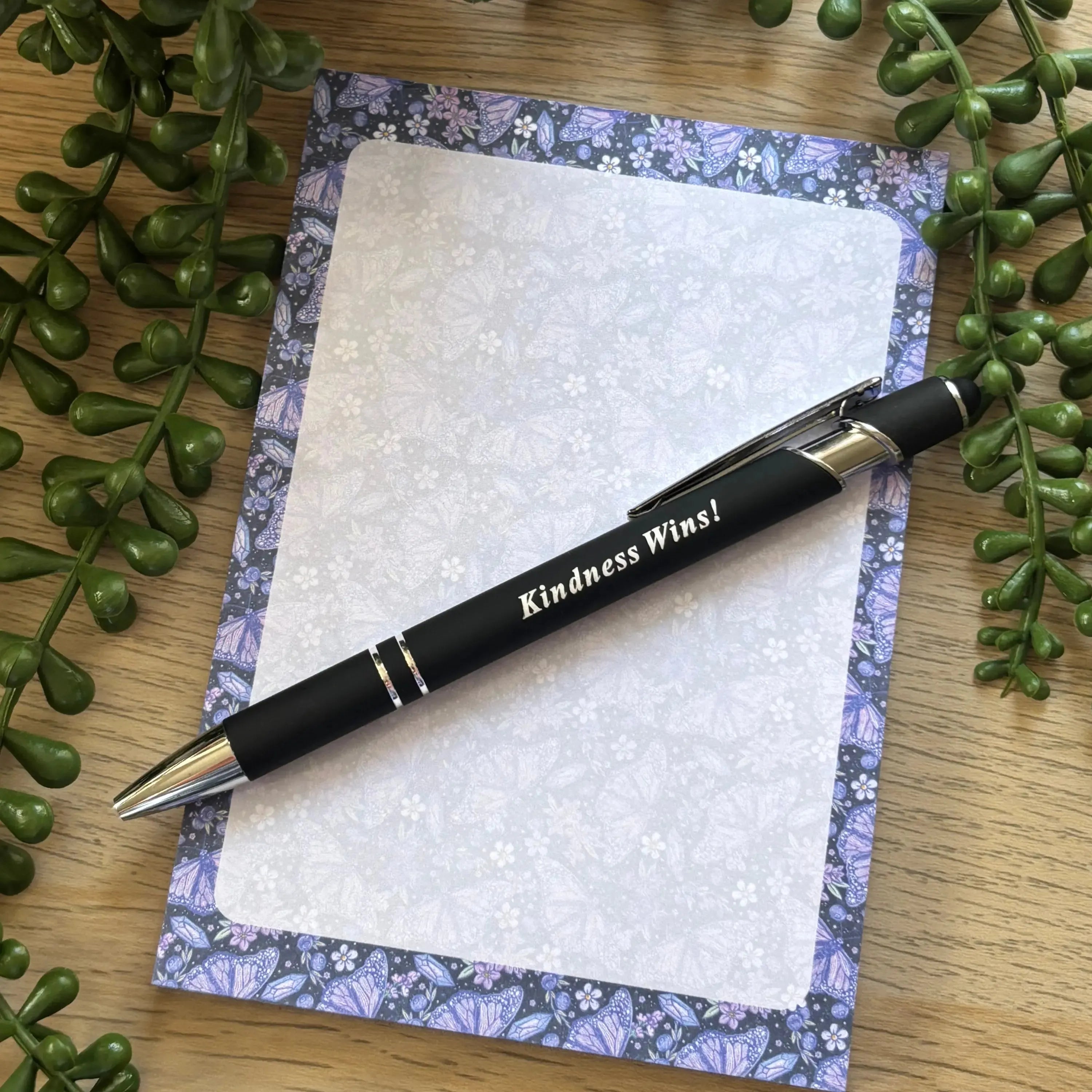 Notepad with purple butterfly border and white floral center, paired with black pen engraved "Kindness Wins!".