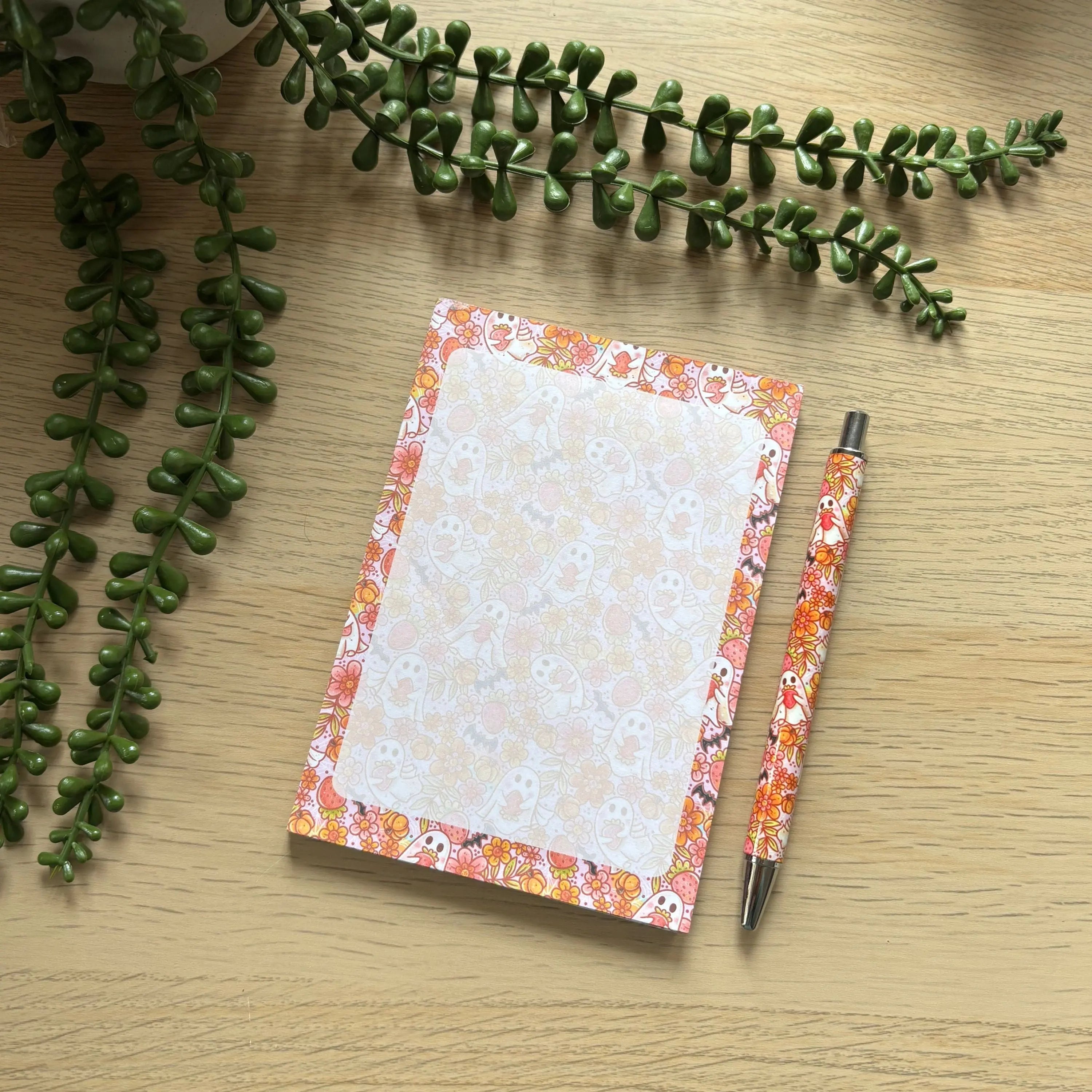 Small Strawberry Ghosts notepad with orange ombre floral border and matching floral-patterned pen on wooden surface.