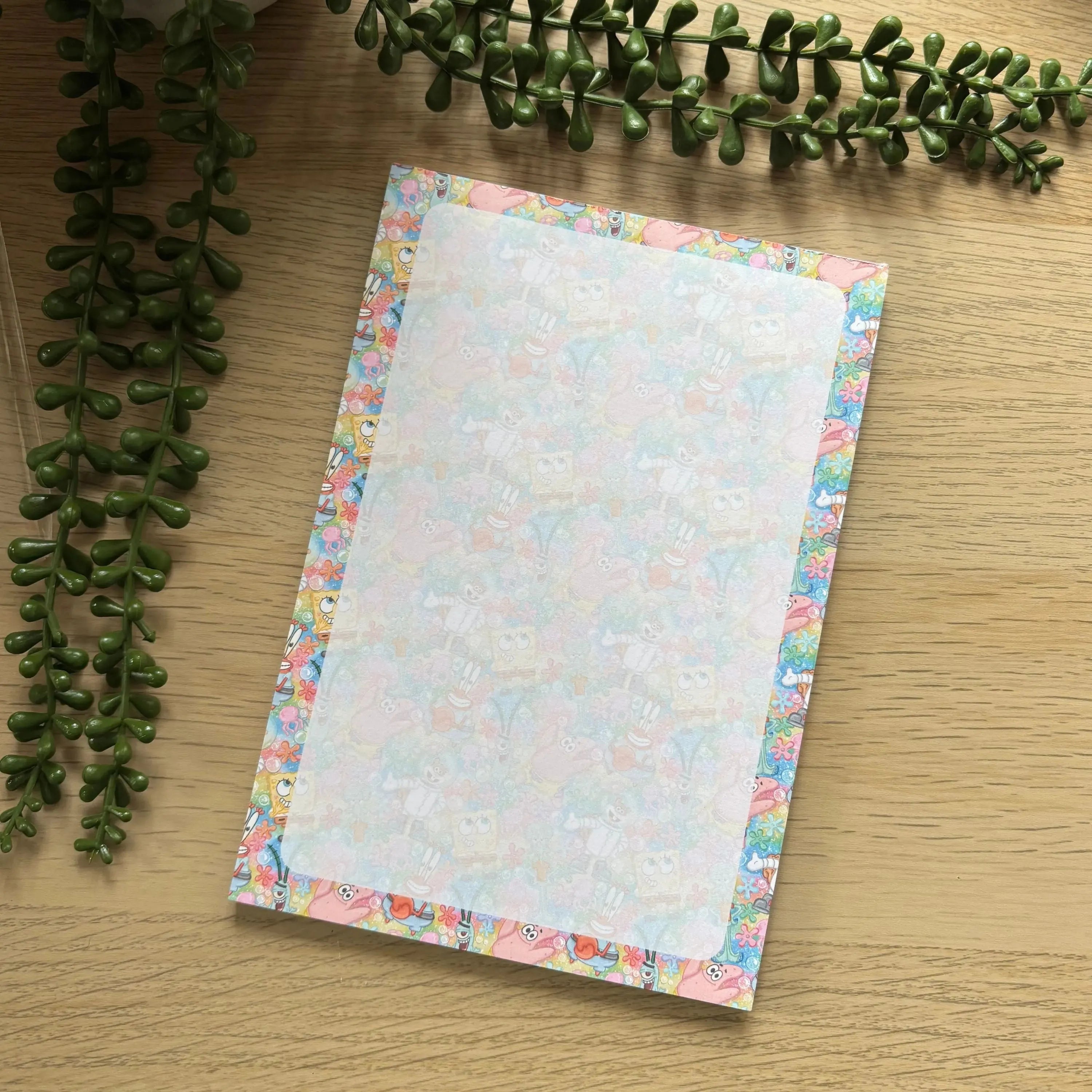 Small notepad with colorful pastel floral border on light wood surface and green foliage nearby.
