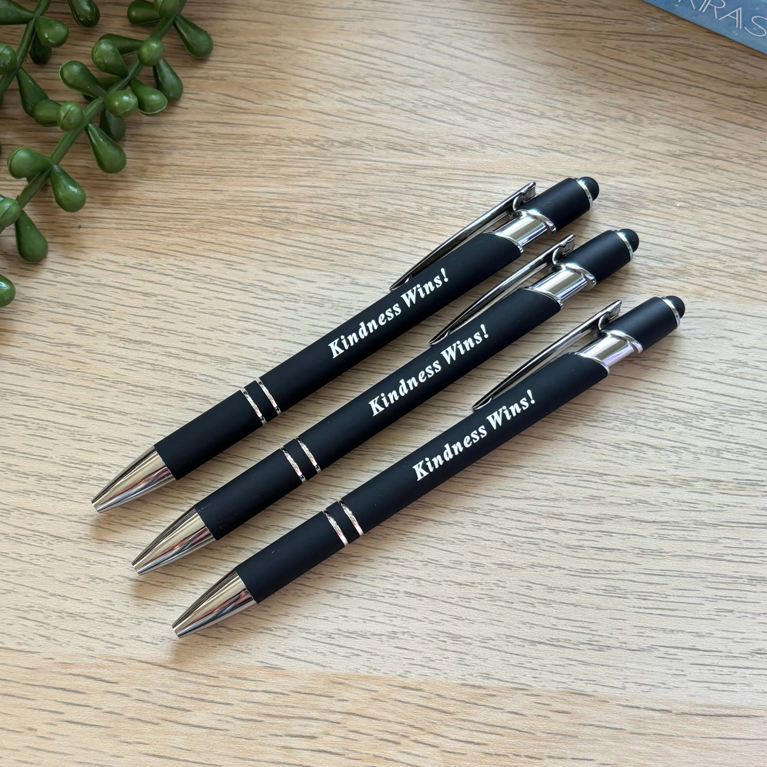 Black metal pen stylus set with "Kindness Wins!" printed in white on a light wood surface.