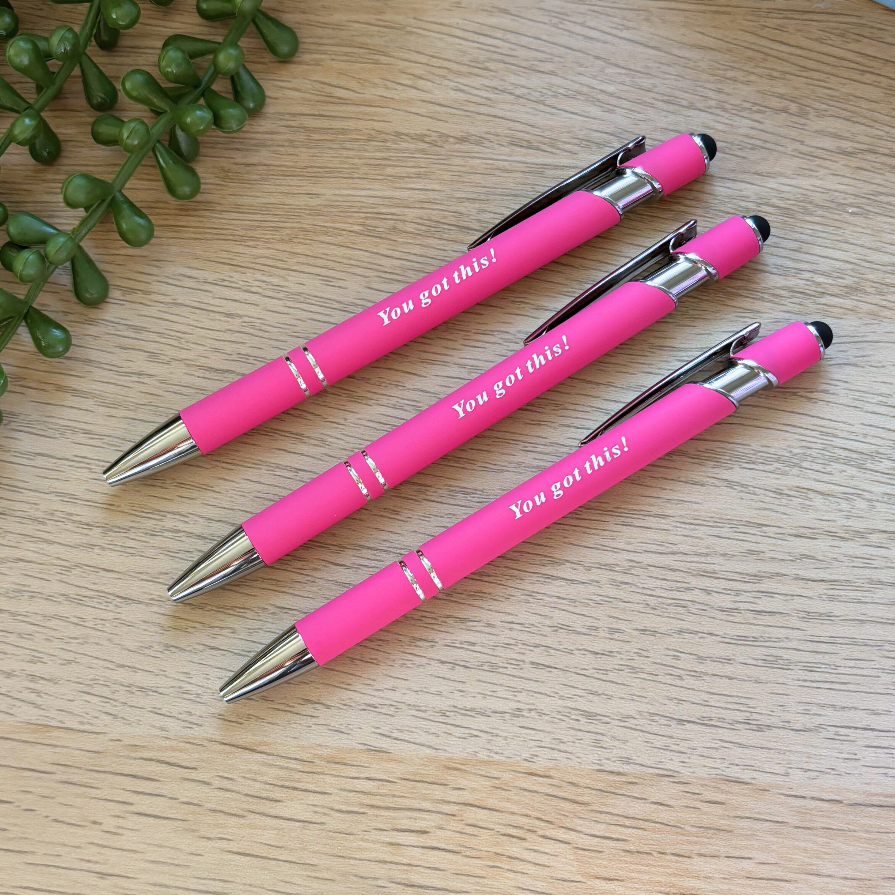 Three bright pink pens with silver tips and clips, each reading "You got this!" in white text, laid on a light wood surface.