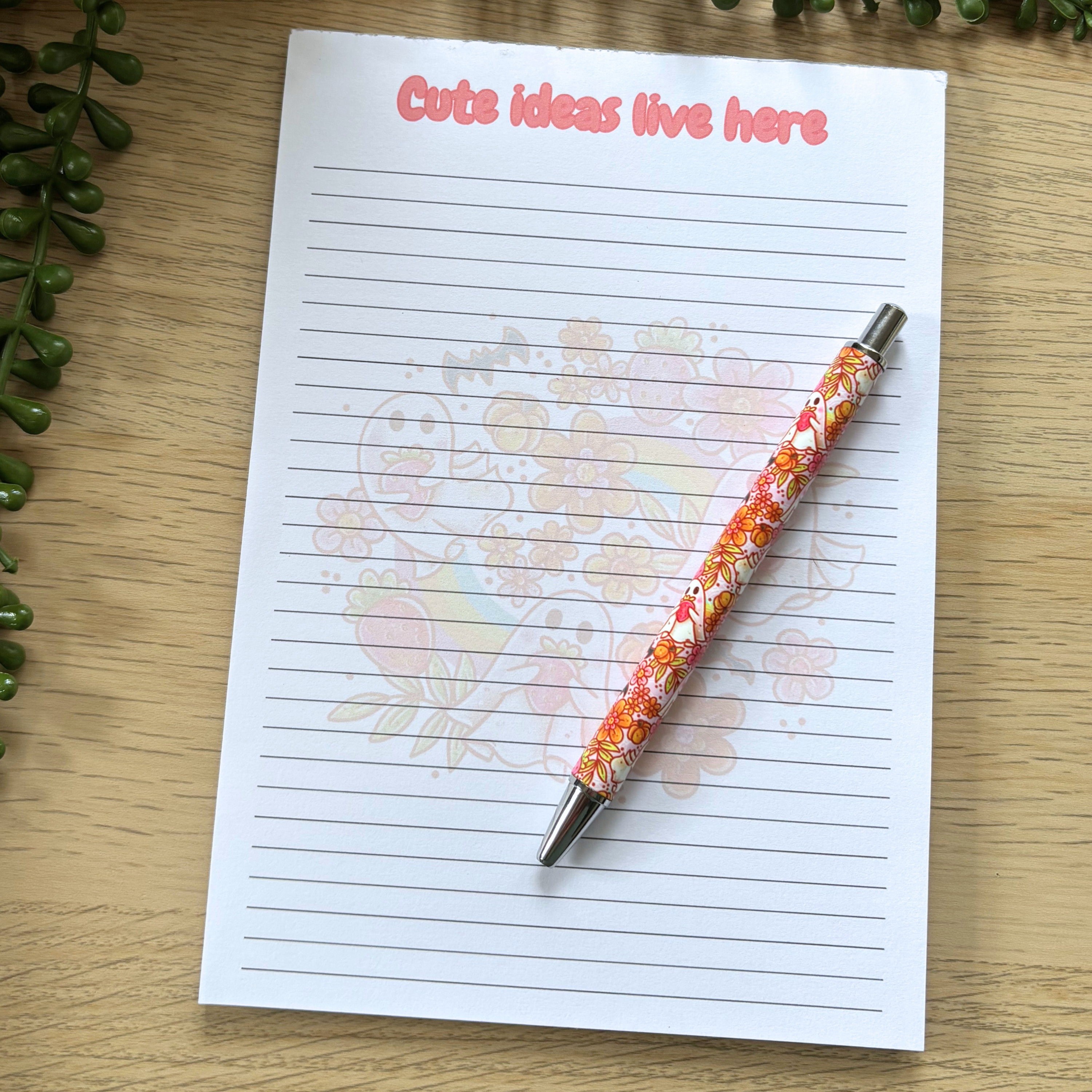 Pen with orange and white strawberry ghost design resting on lined notepad with "Cute ideas live here" text in pink.