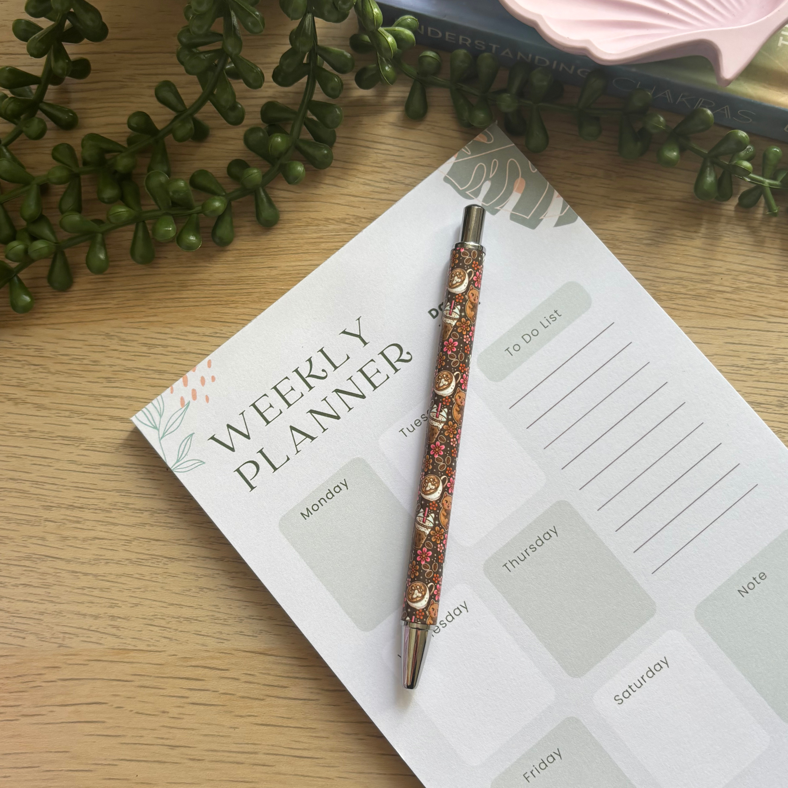 Brown ballpoint pen with coffee cup and cat illustrations resting on a weekly planner on a wooden surface.