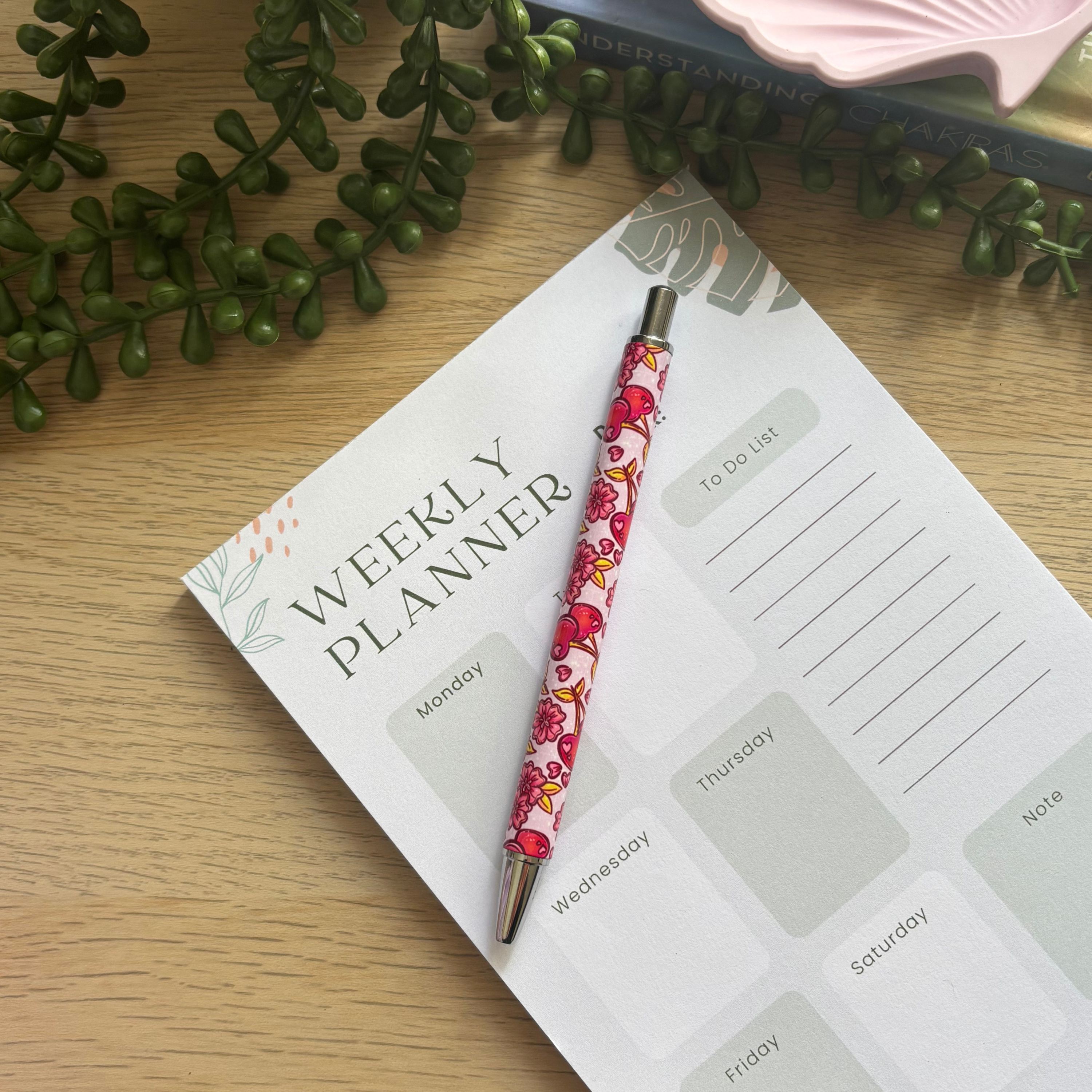 Pink cherry blossom patterned pen resting on a weekly planner page with green leafy decor in the background.