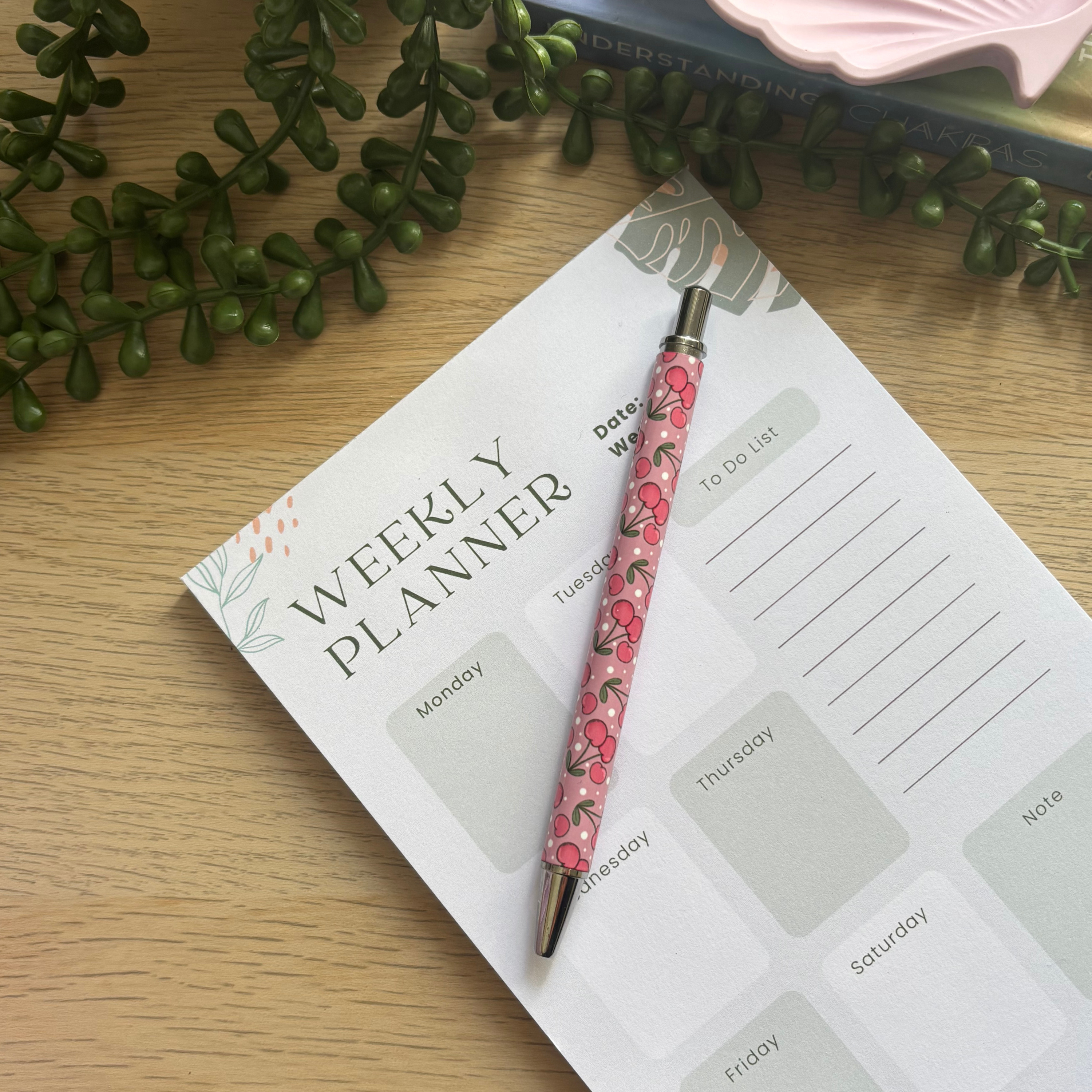Pink pen with red cherry patterns resting on a weekly planner page with green plant decor nearby.