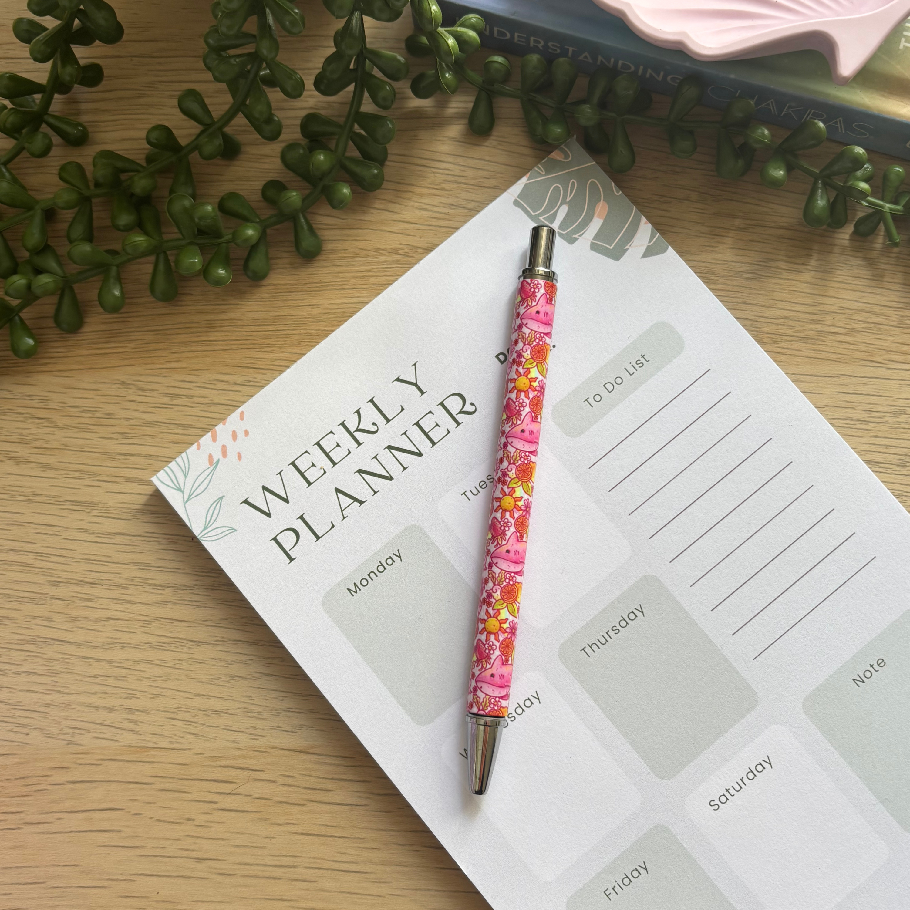 Pink floral lemon shark patterned ballpoint pen resting on a weekly planner with green plant decoration in the background.