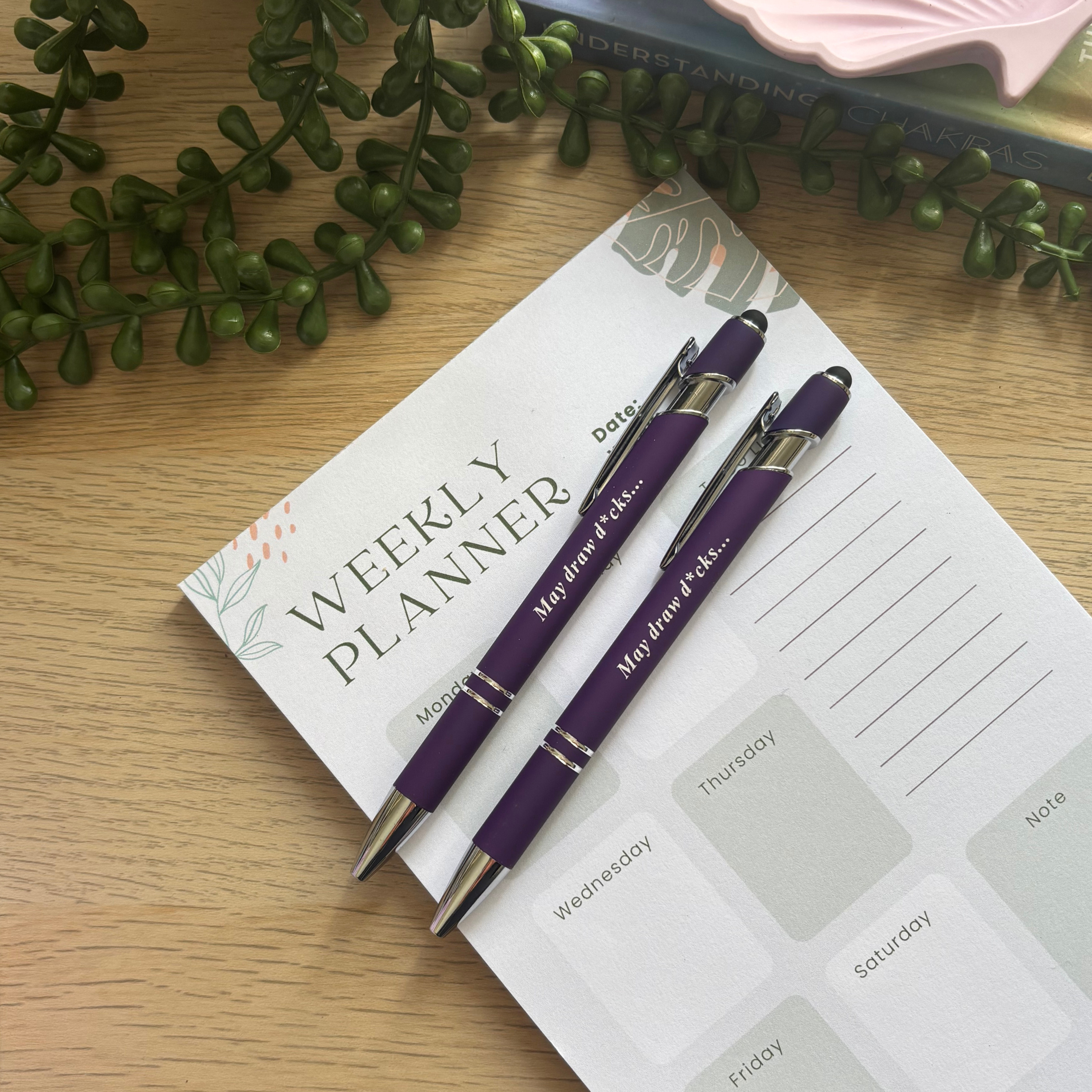 Two purple ballpoint pens with silver accents and humorous text on a weekly planner page.