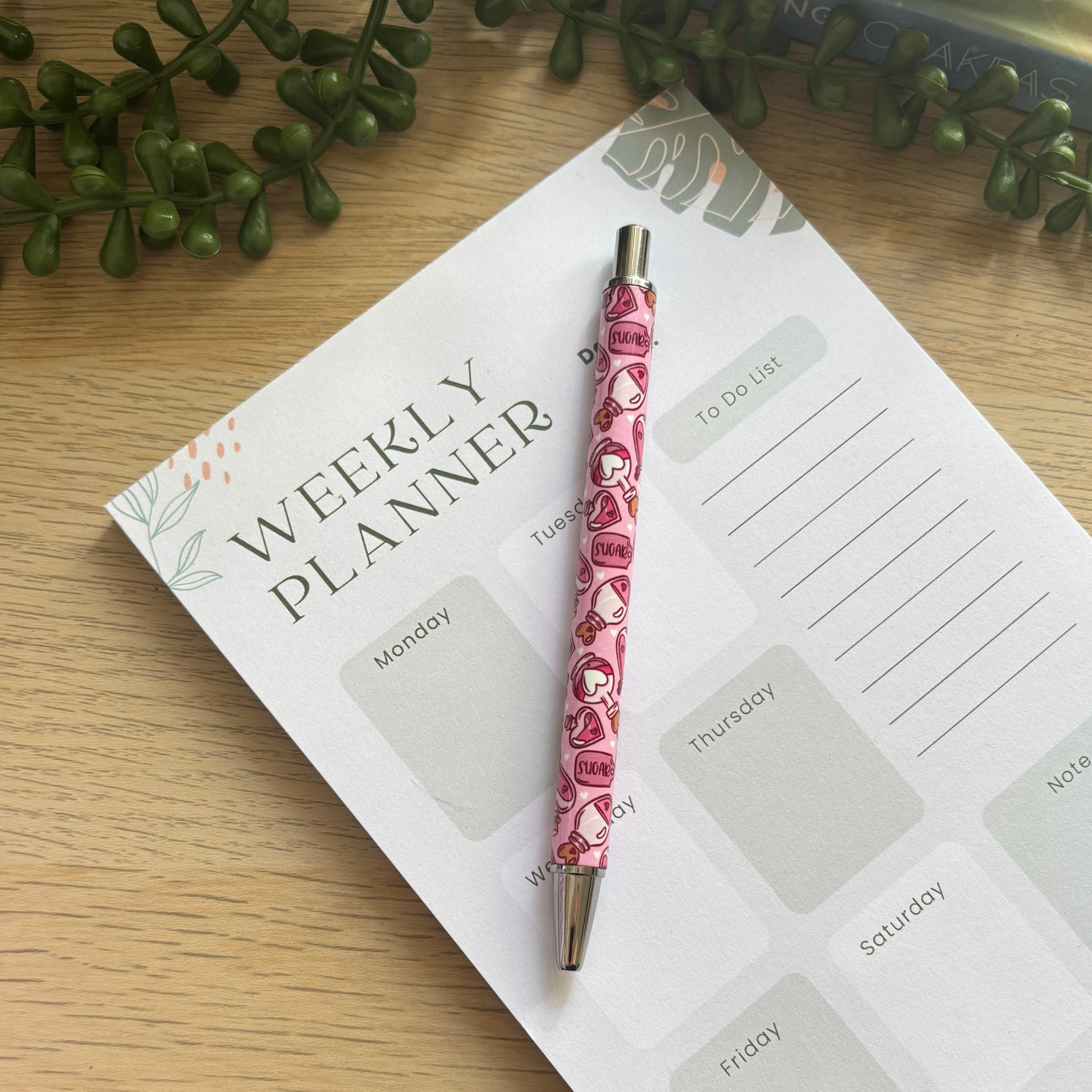 Pink pen with heart and "sugar and spice" text pattern resting on weekly planner with leafy design.