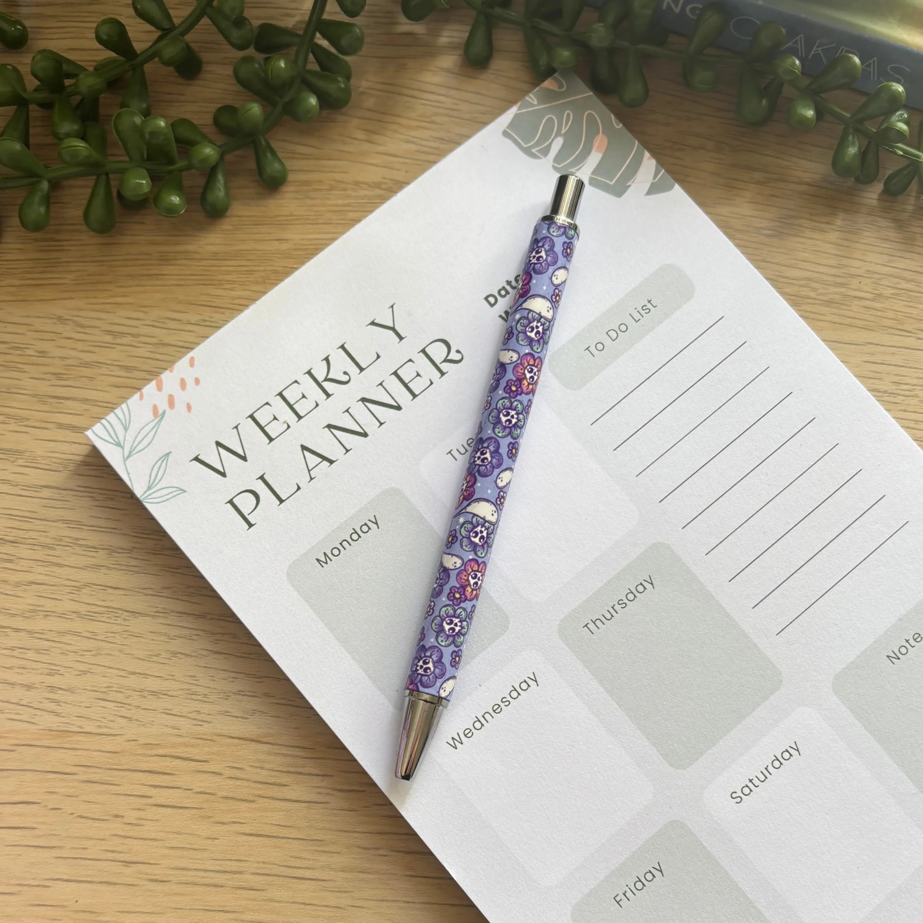 Purple ballpoint pen with small ghost and flower pattern resting on a weekly planner pad.