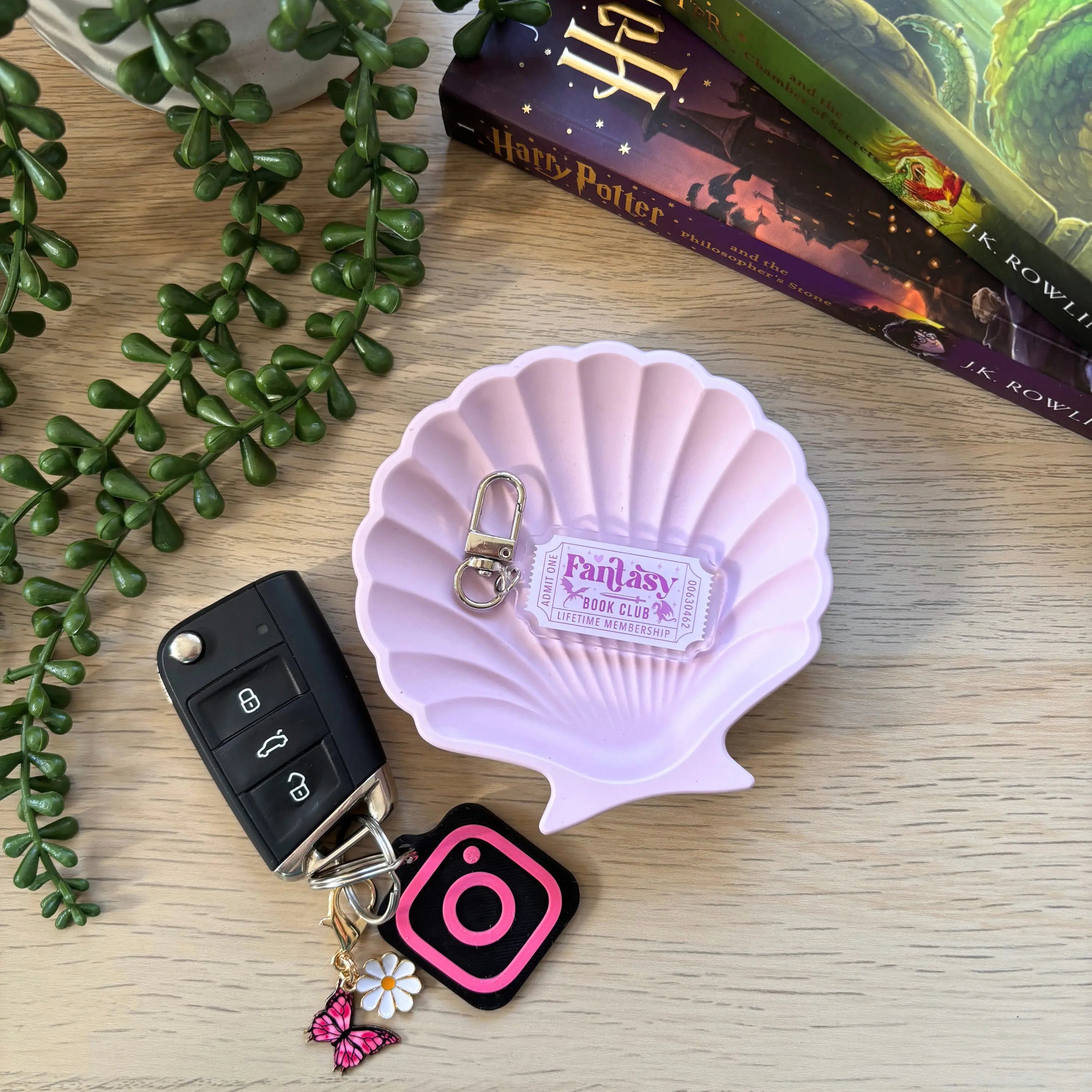 Pink seashell-shaped tray holding a Fantasy Bookish Ticket keyring with butterfly and flower charms on a wooden surface next .