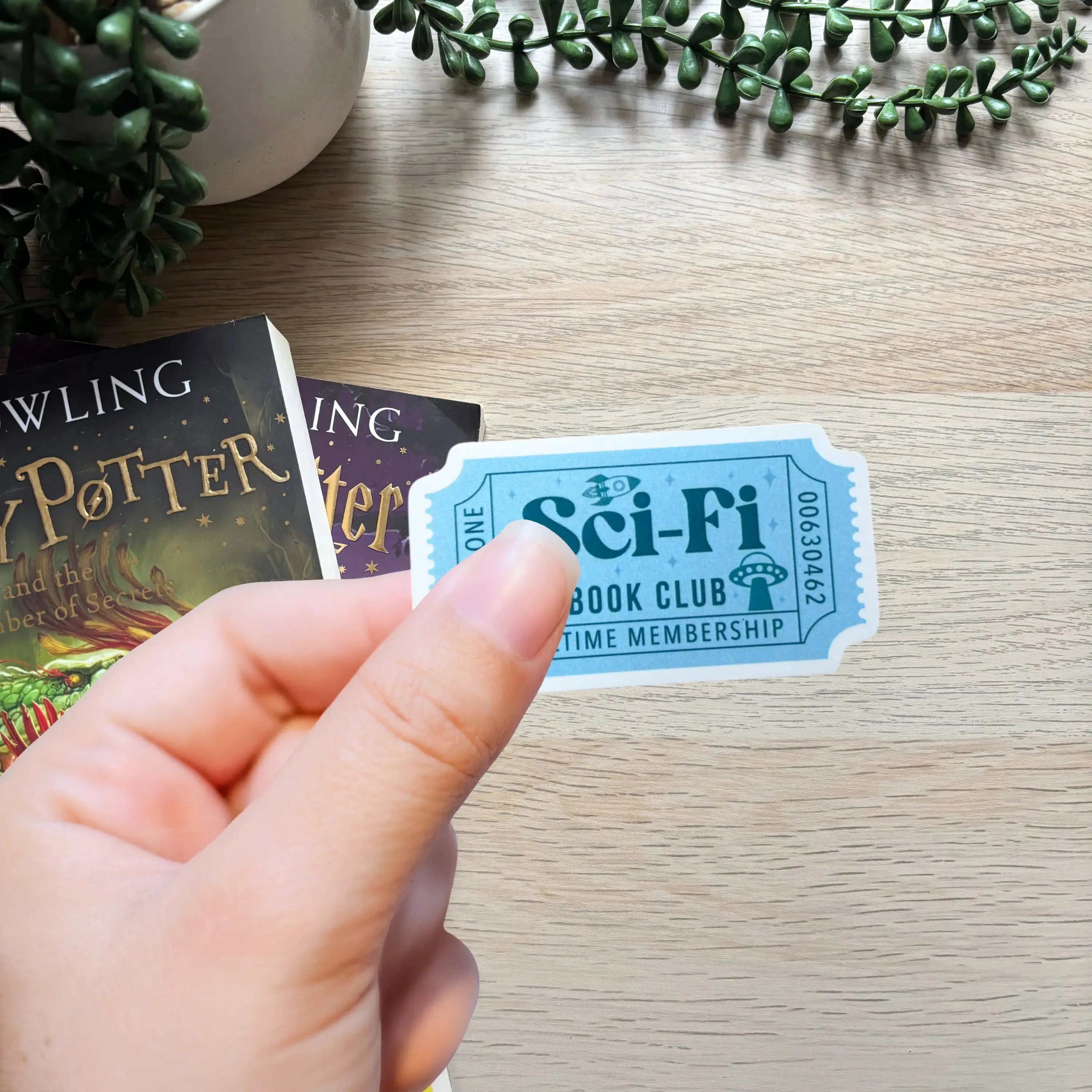 Blue rectangular sticker with vintage ticket design reading "Sci-Fi Book Club Lifetime Membership" held over a wood surface.