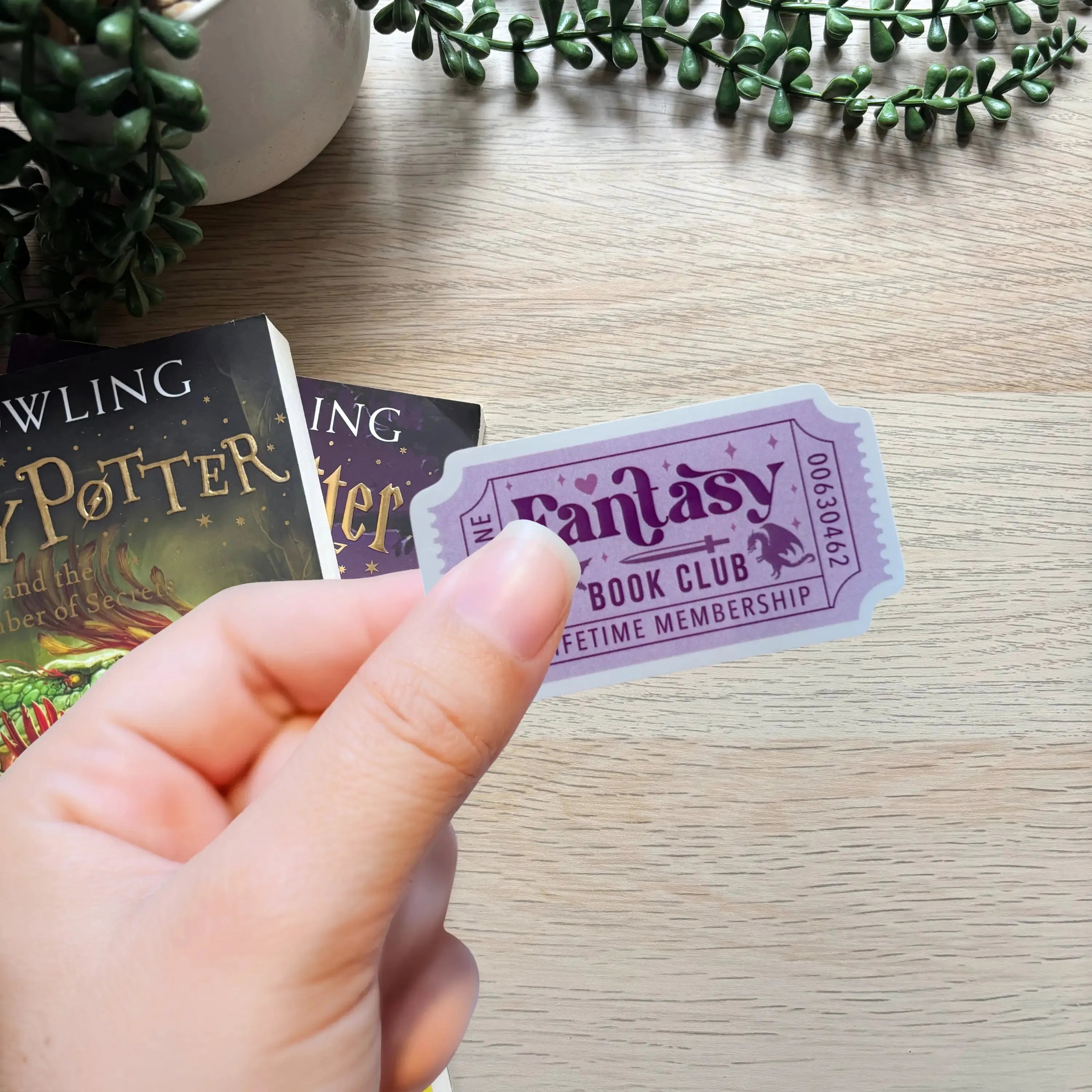 Purple ticket-shaped sticker with "Fantasy Book Club" and decorative stars held over a wooden surface.