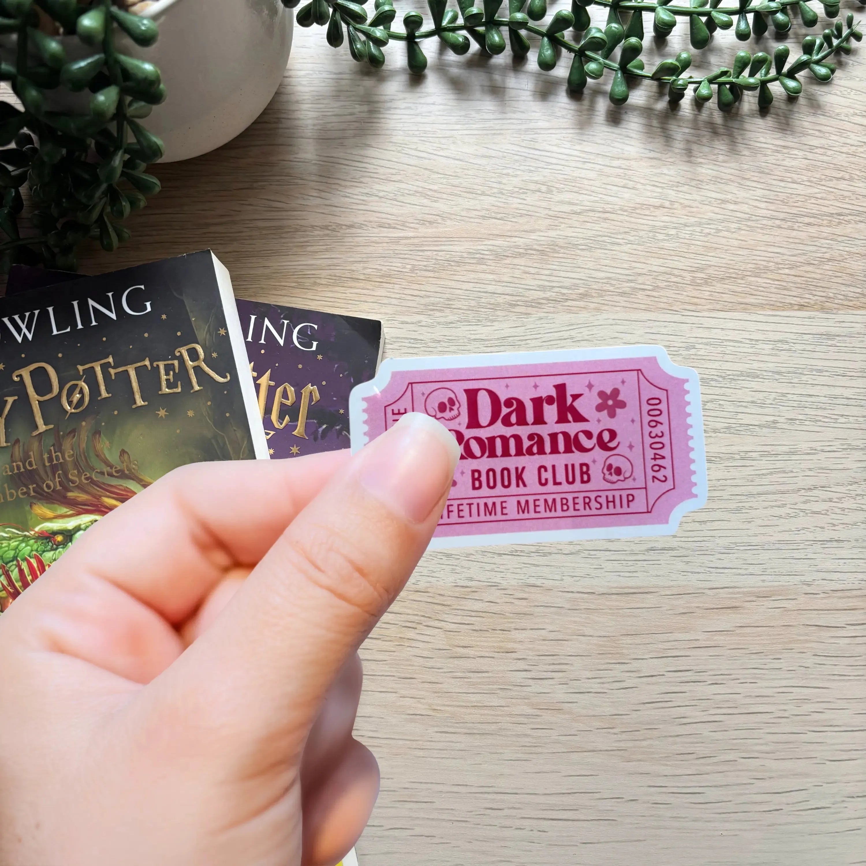 Pink "Dark Romance Book Club Lifetime Membership" sticker with decorative border held by hand over wooden surface with books .