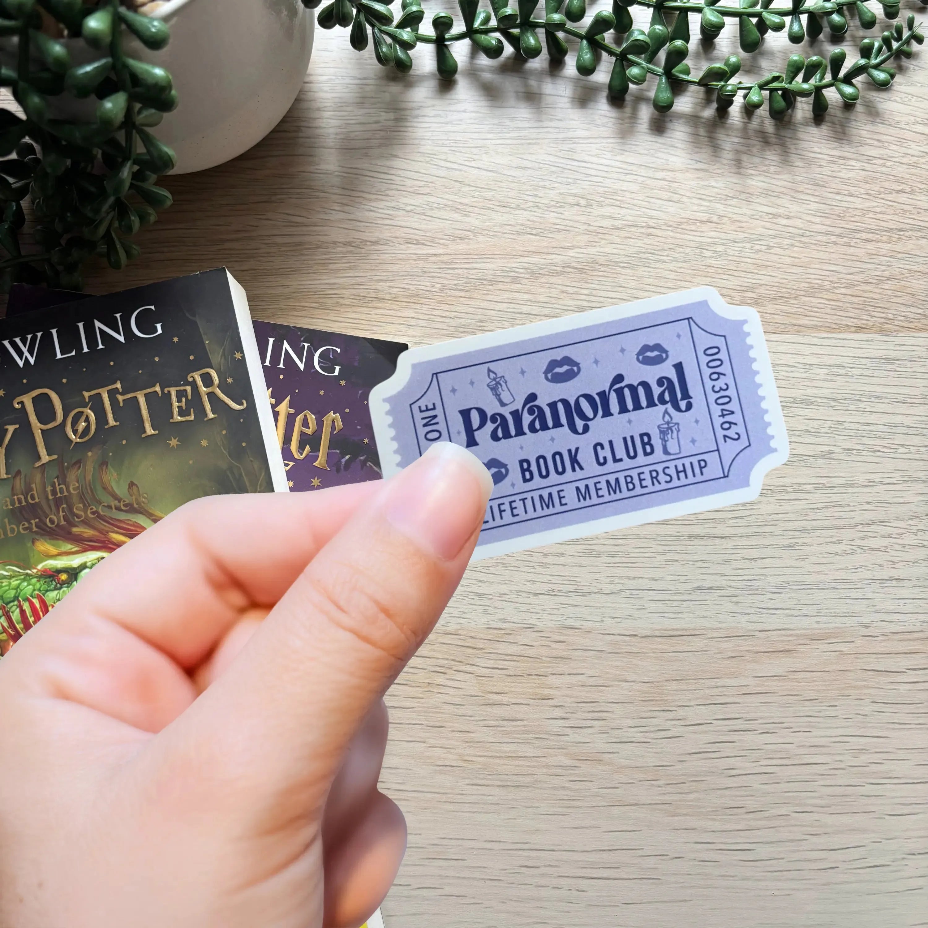 Hand holding a white sticker shaped like a ticket with blue text reading Paranormal Book Club Lifetime Membership.