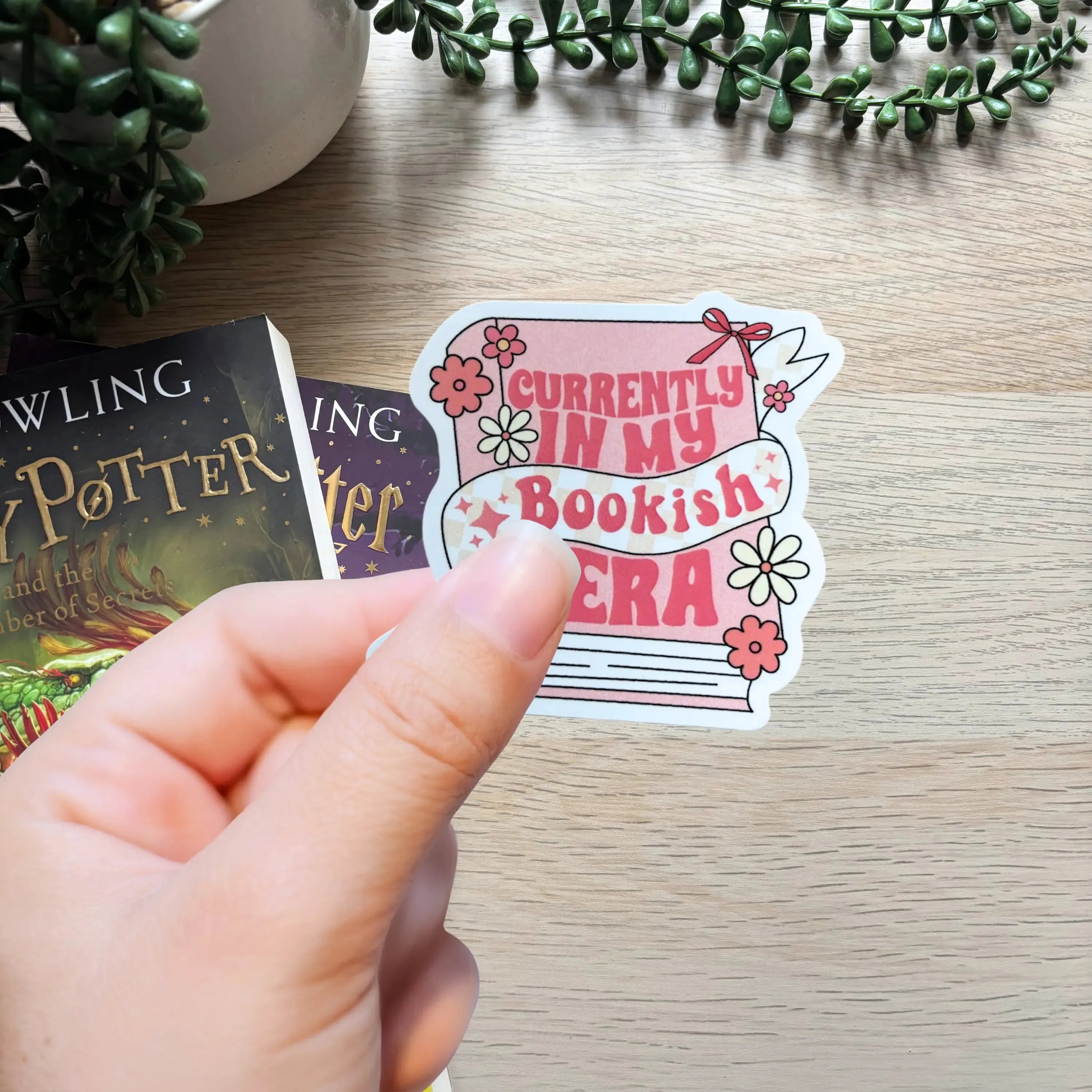 Hand holding a pink and white sticker with "Currently in my Bookish Era" text and floral design on wooden surface next to.