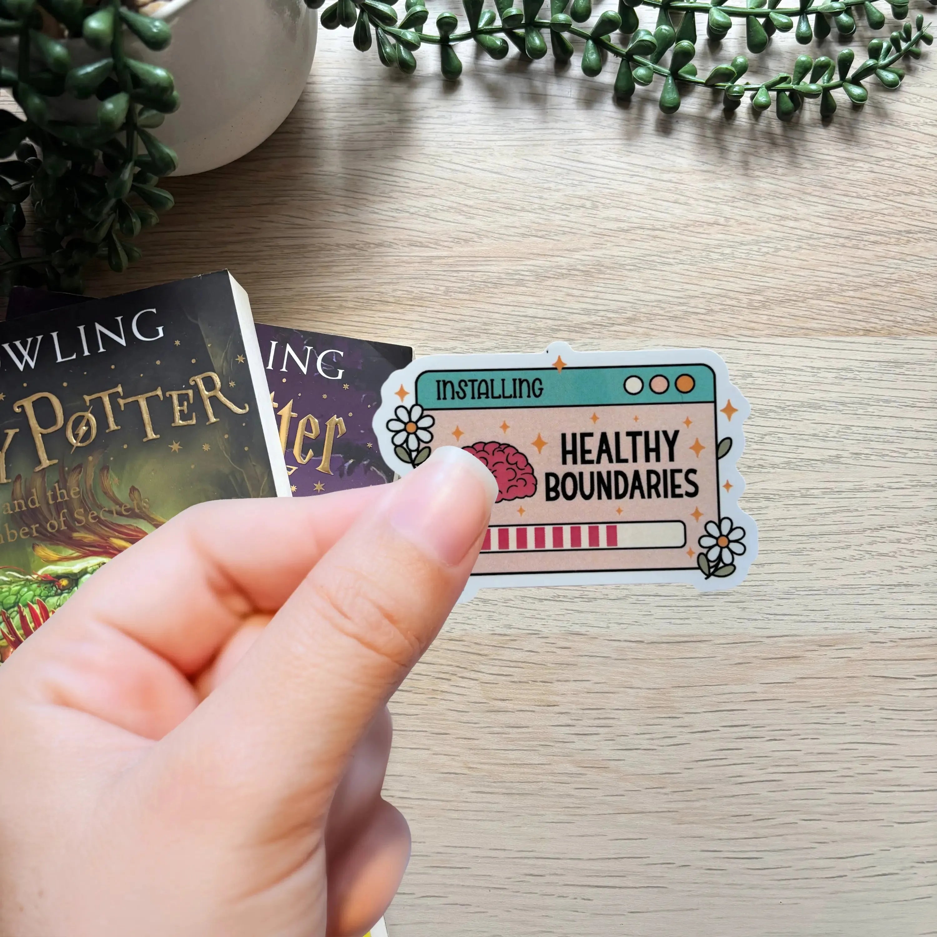 Hand holding colorful Installing Healthy Boundaries sticker with progress bar and floral accents on wooden surface.