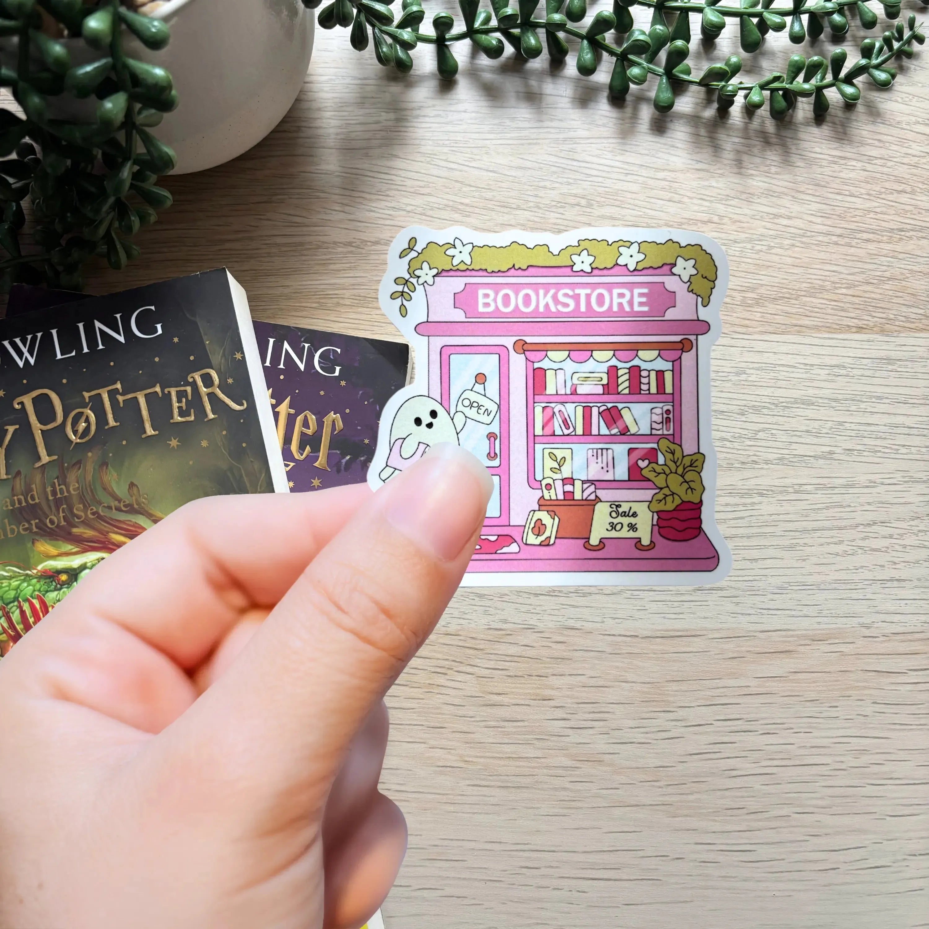 Hand holding colorful sticker of a spooky pink bookstore with ghost and pumpkins on wooden table.