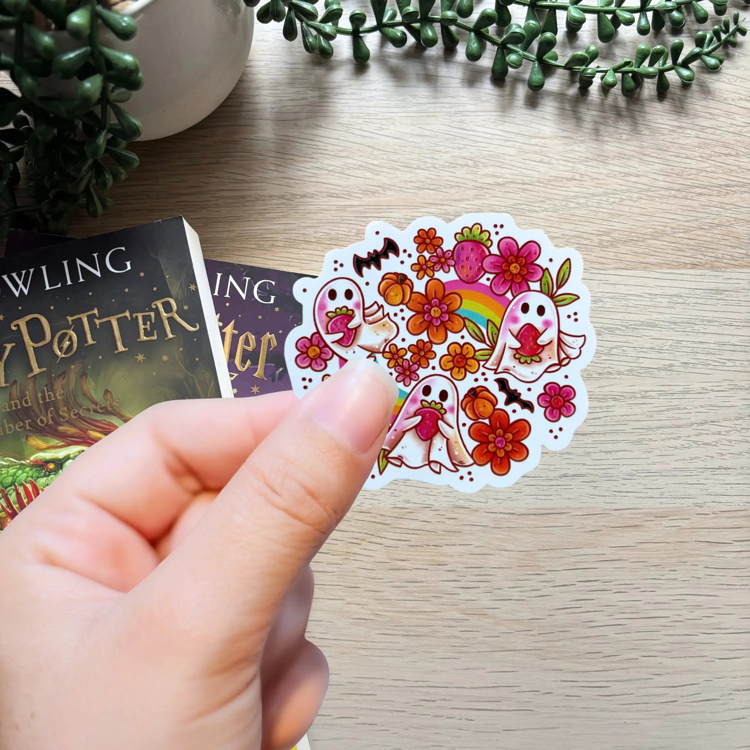 Hand holding colorful sticker with cute white ghosts, strawberries, and flowers.
