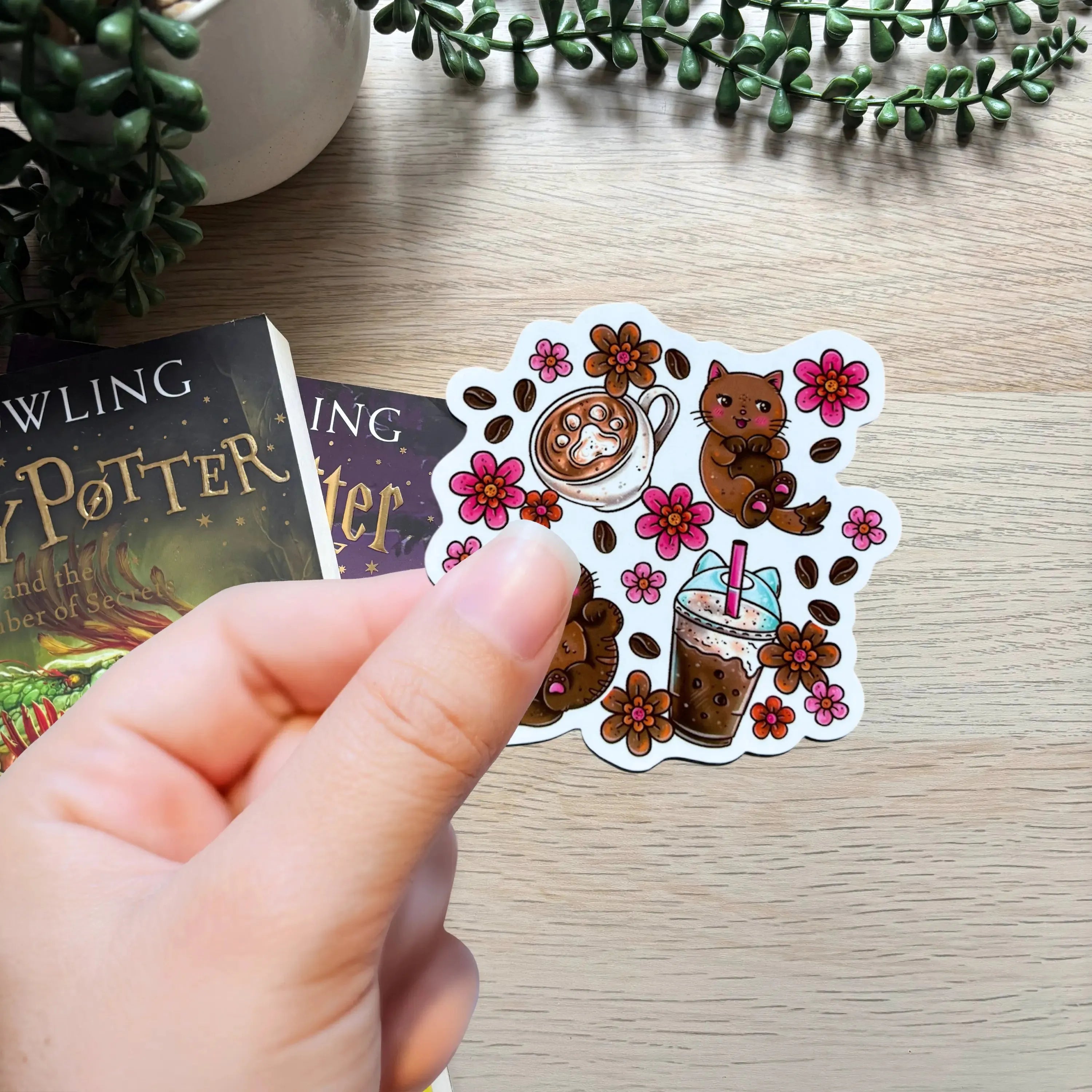 Hand holding Coffee Cats sticker with brown cats, coffee cups, and pink flowers on wood table.
