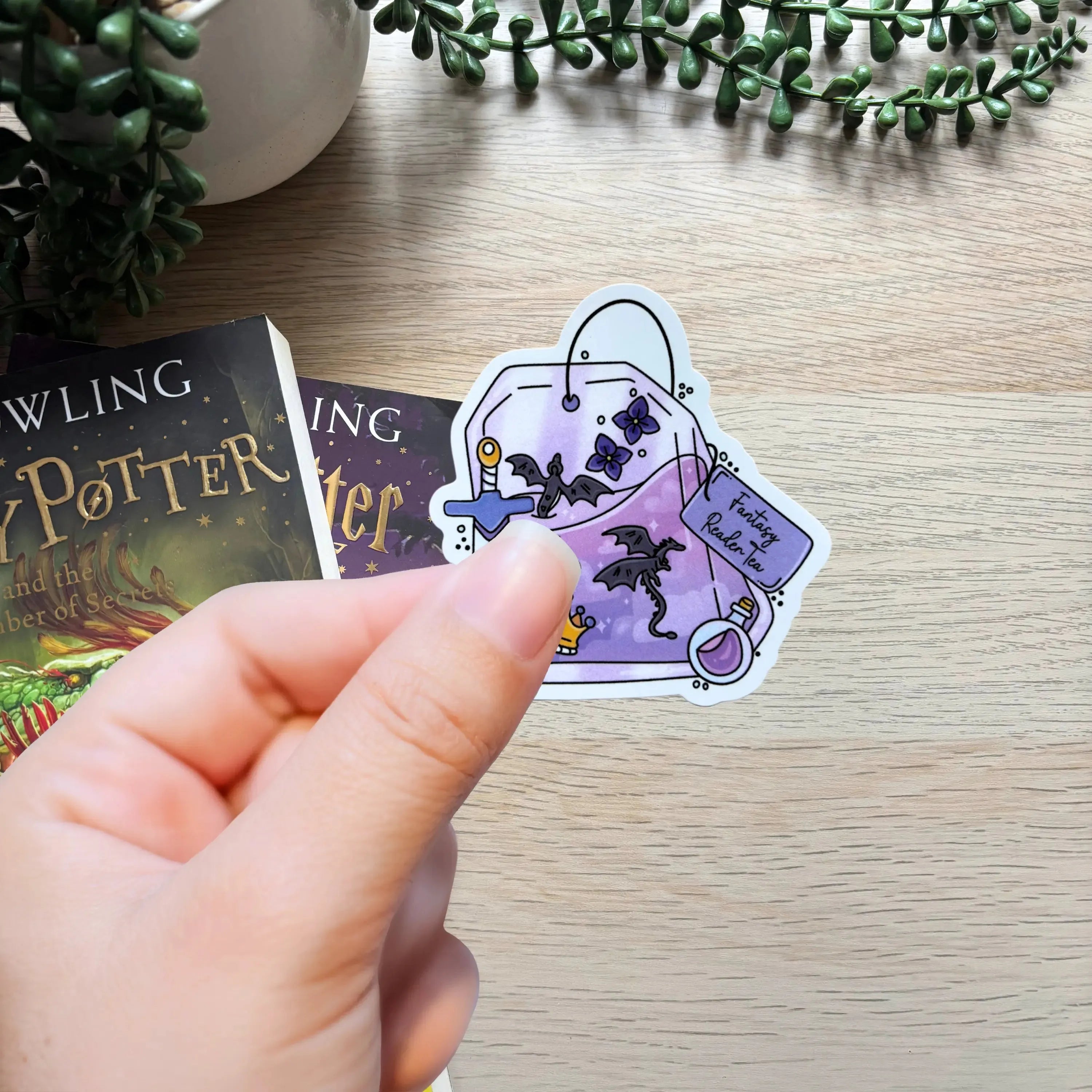 Purple fantasy-themed sticker with books, sword, key, and magical symbols held above wooden table.