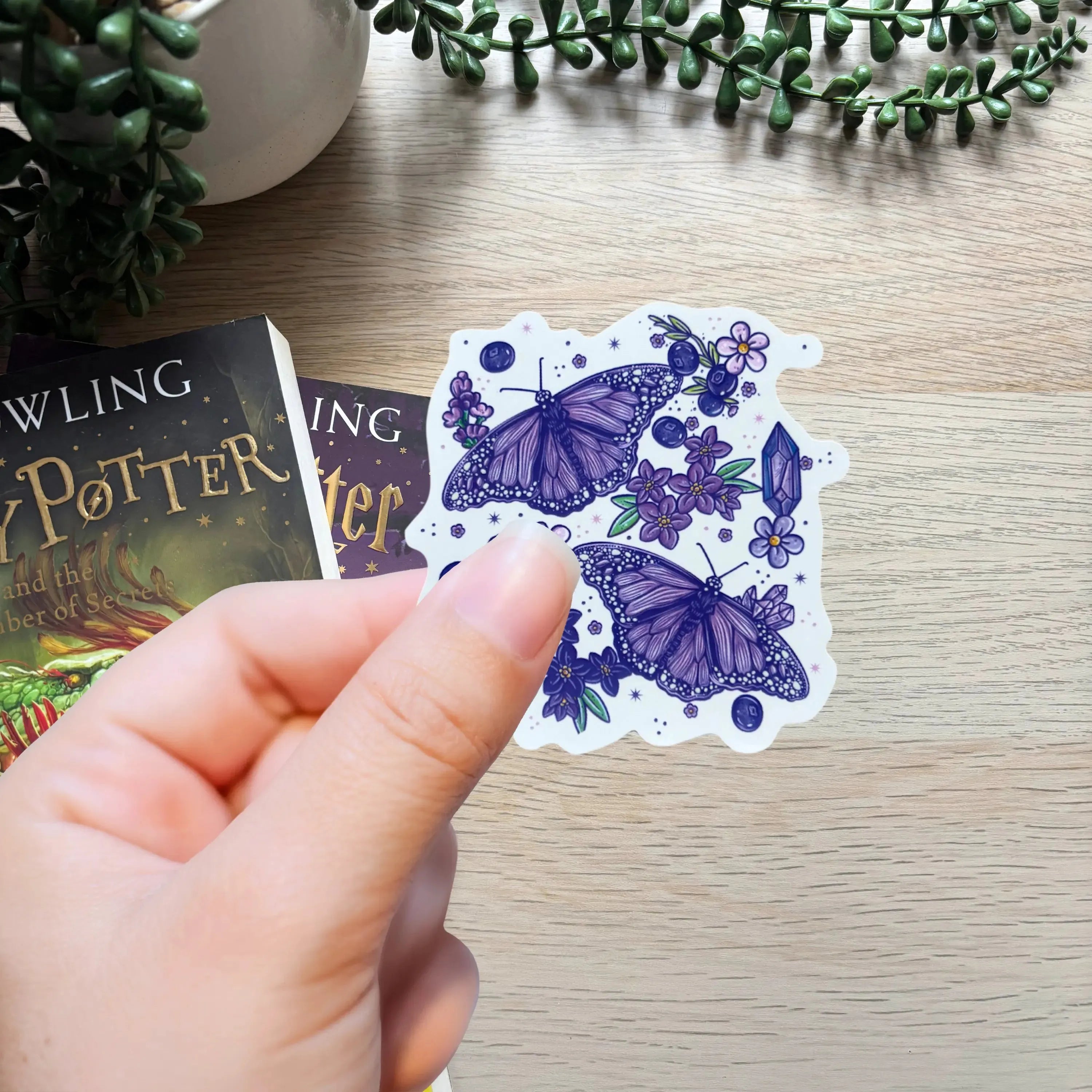 Hand holding a card with purple butterfly design next to a 'Harry Potter' book on a wooden surface.