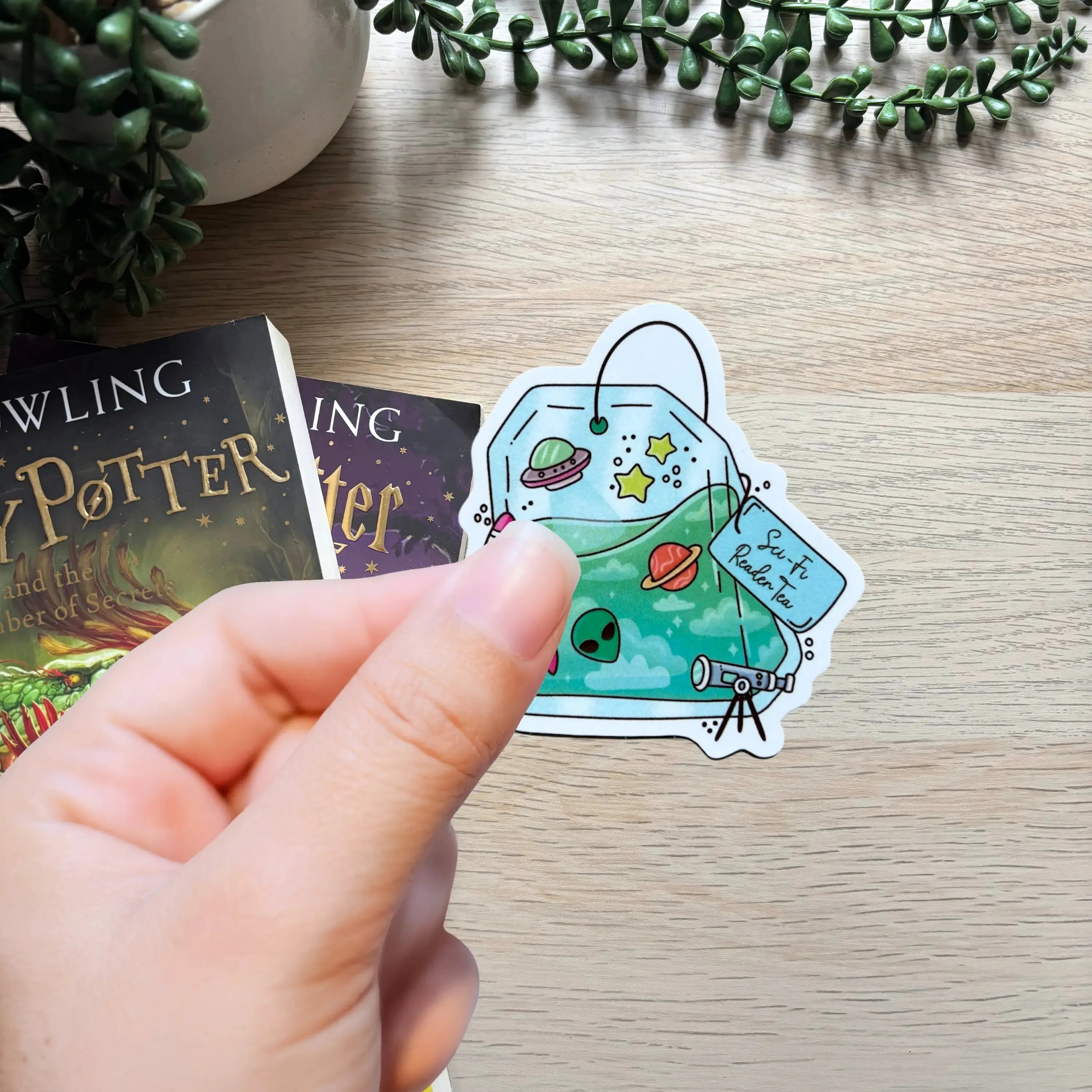 Hand holding a colorful sci-fi themed sticker with planets, stars, and a book on a wooden table.