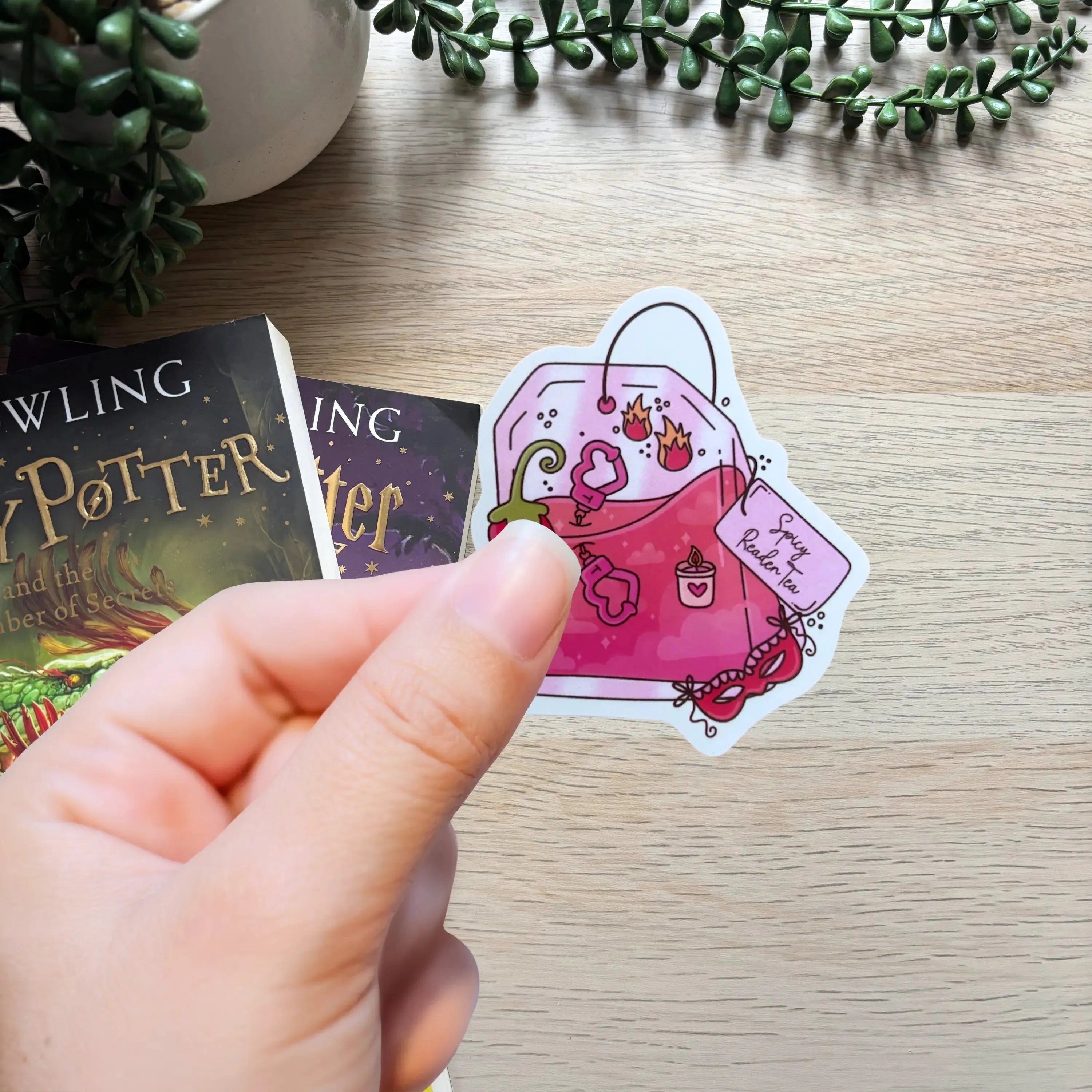 Hand holding a colorful sticker of a pink handbag filled with books and a heart tag on wood surface.