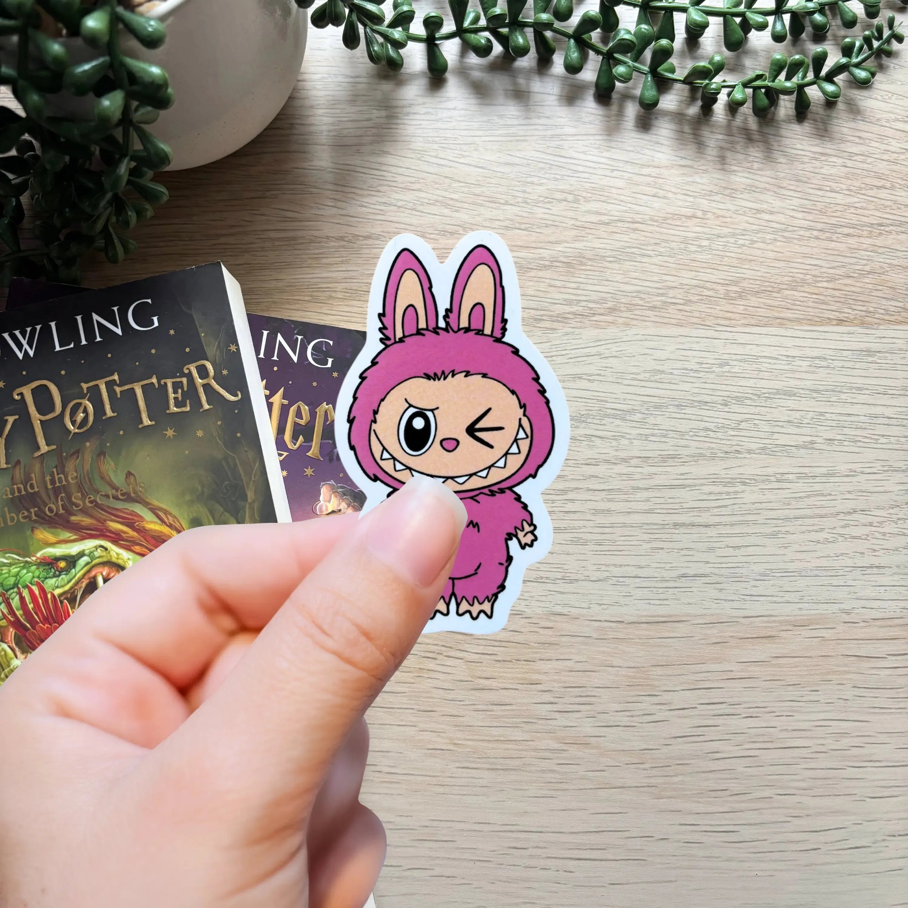 Hand holding a small pink cartoon bunny sticker winking on a wooden surface next to Harry Potter books.