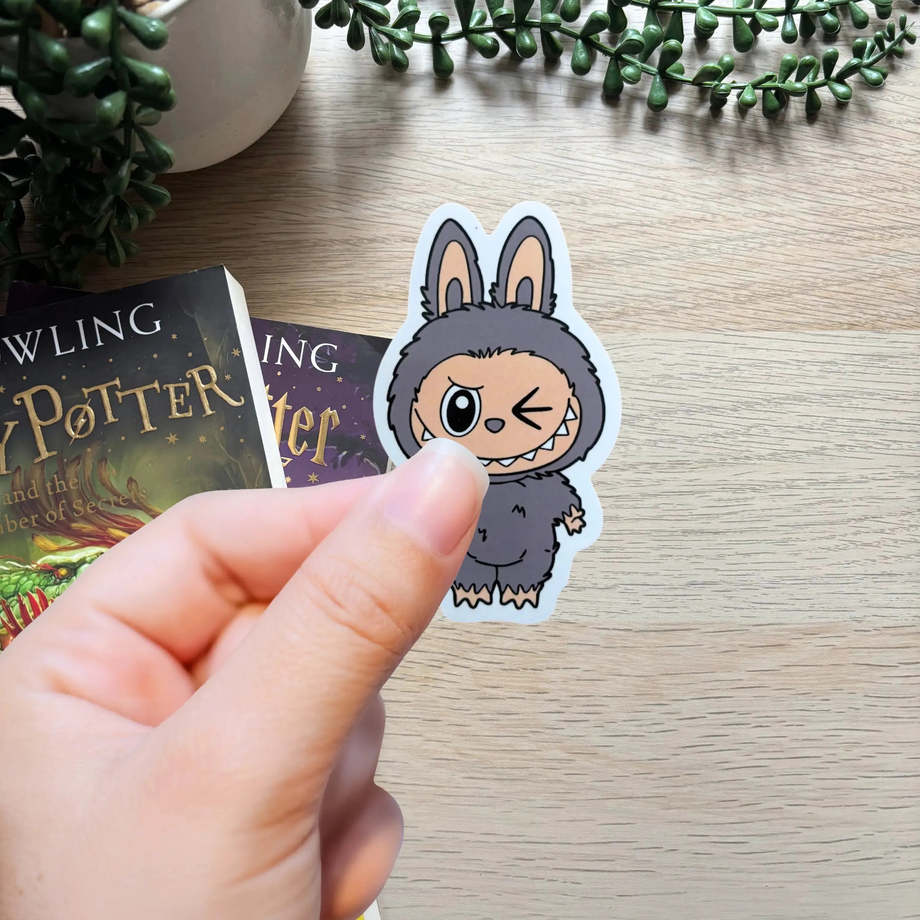 Hand holding small grey bunny character sticker with big eyes and floppy ears on wooden table background.