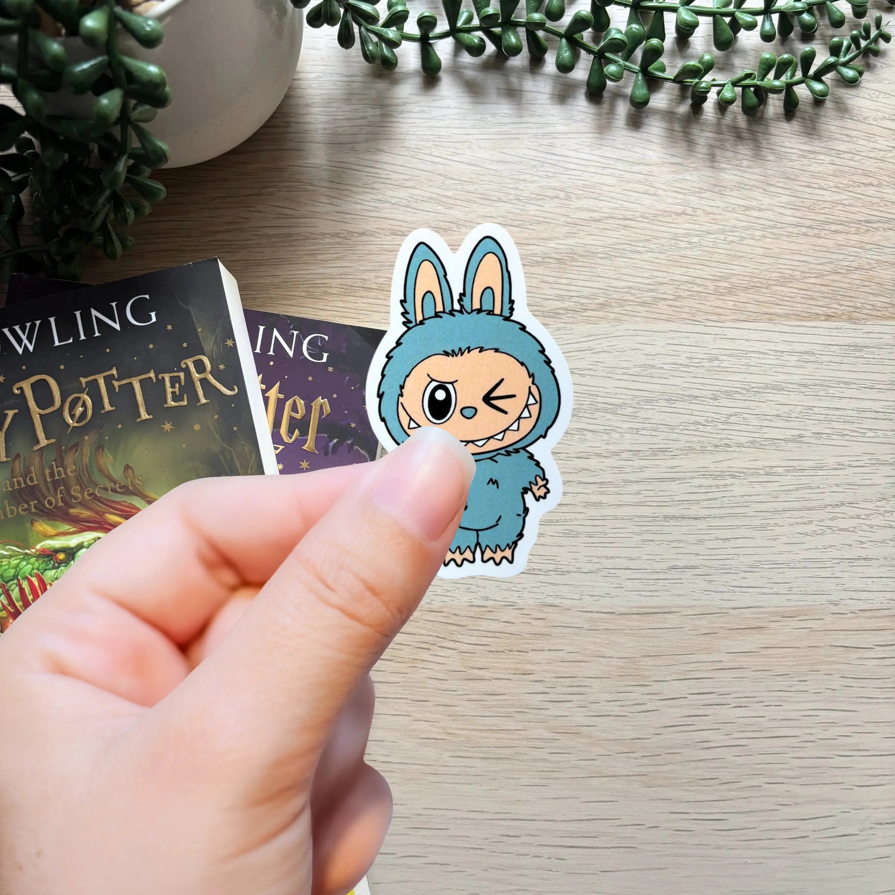 Hand holding a small blue bunny character sticker winking on light wood surface with books and greenery nearby.
