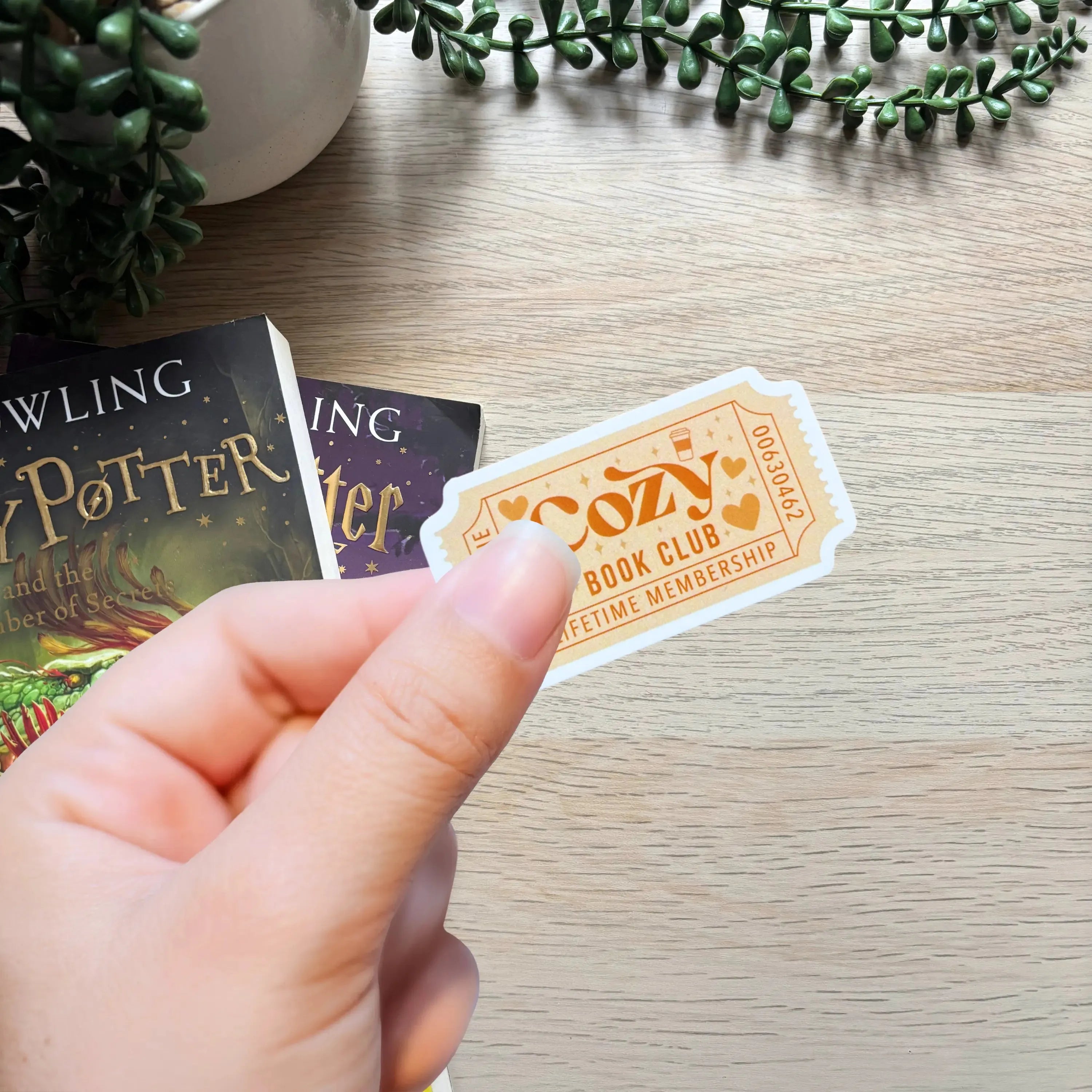 Hand holding a beige and orange Cozy Book Club sticker with decorative font and border on a light wood surface with Harry.