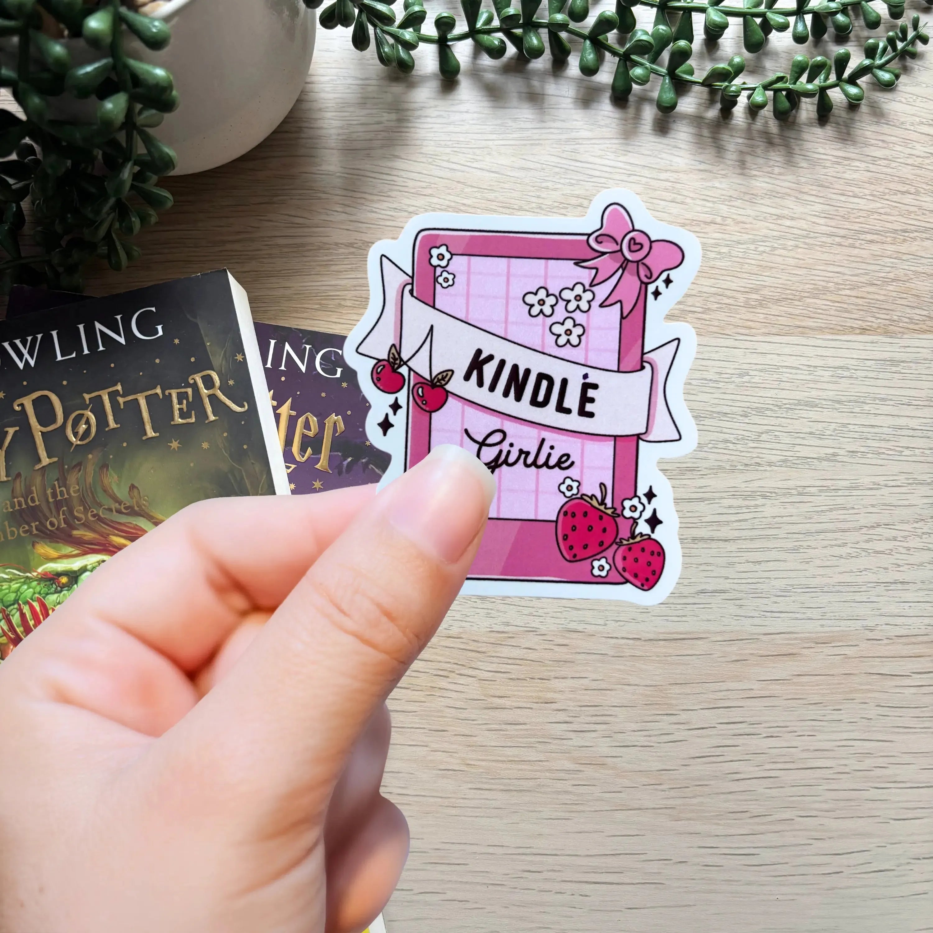 Hand holding pink Kindle-themed sticker with book, flower, and strawberry illustrations on wooden surface.