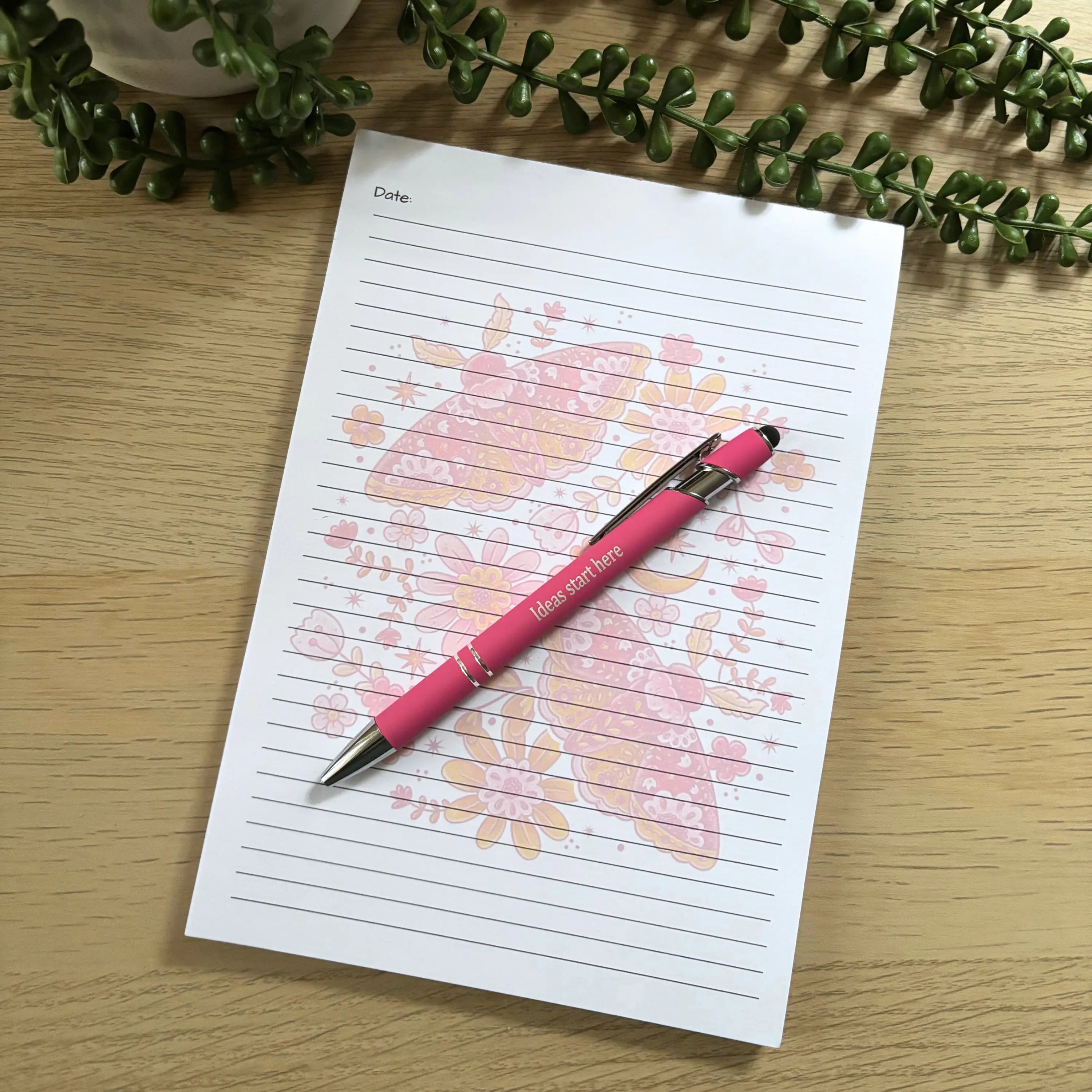 A5 lined notepad with a pink moth and floral design and a matching pink pen on wooden surface.