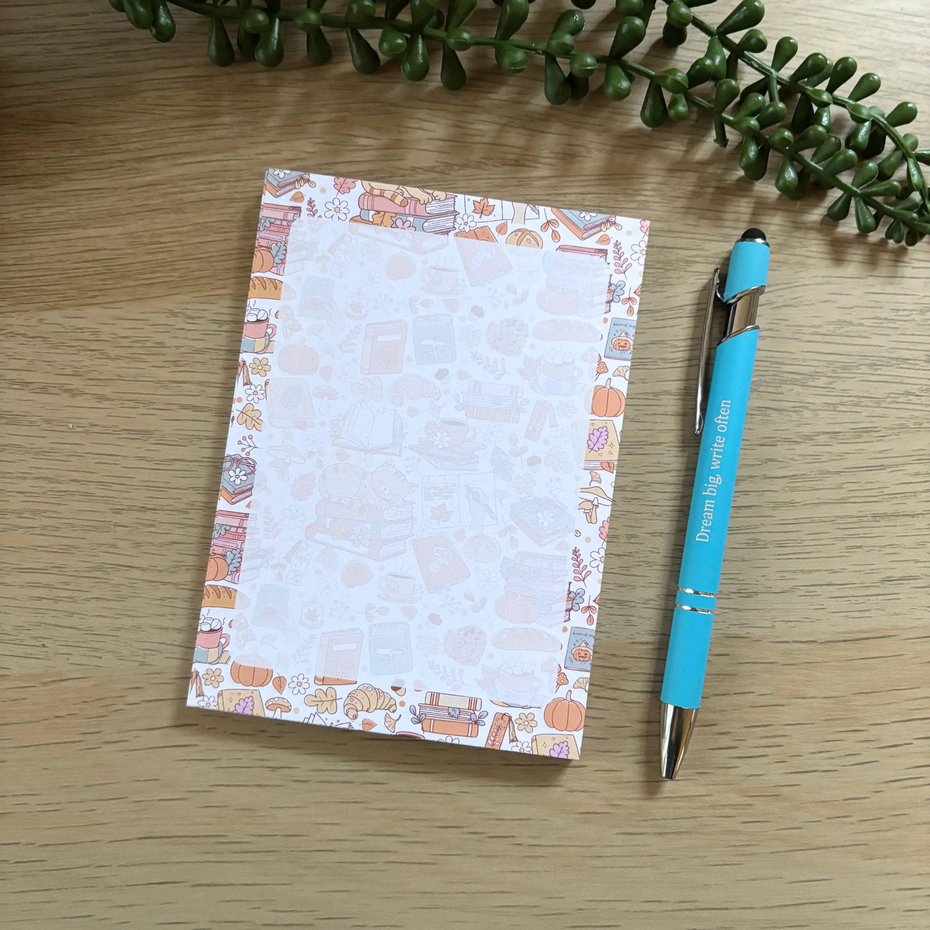 Small cozy autumn cat notepad with orange cat illustrations and a turquoise pen on wood table.