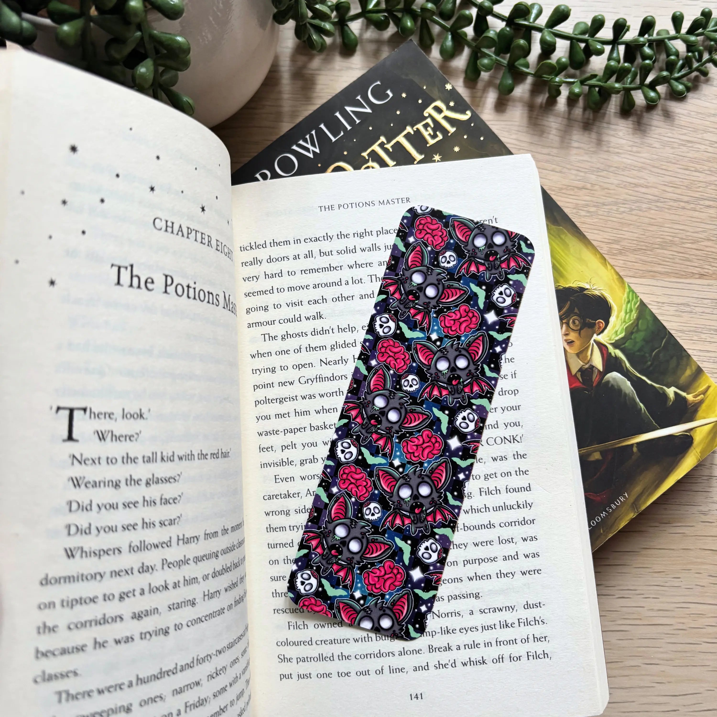 Black bookmark with colorful zombie bats, skulls, and roses pattern, placed in an open book on a wooden table.