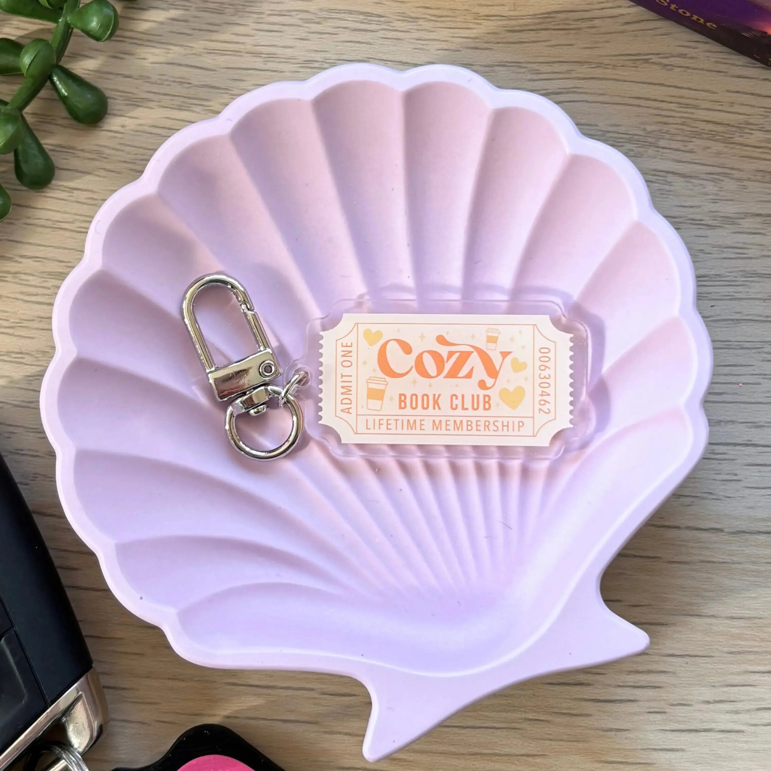 Pink and beige Cozy Bookclub keyring with metal clasp on a lavender shell-shaped dish.