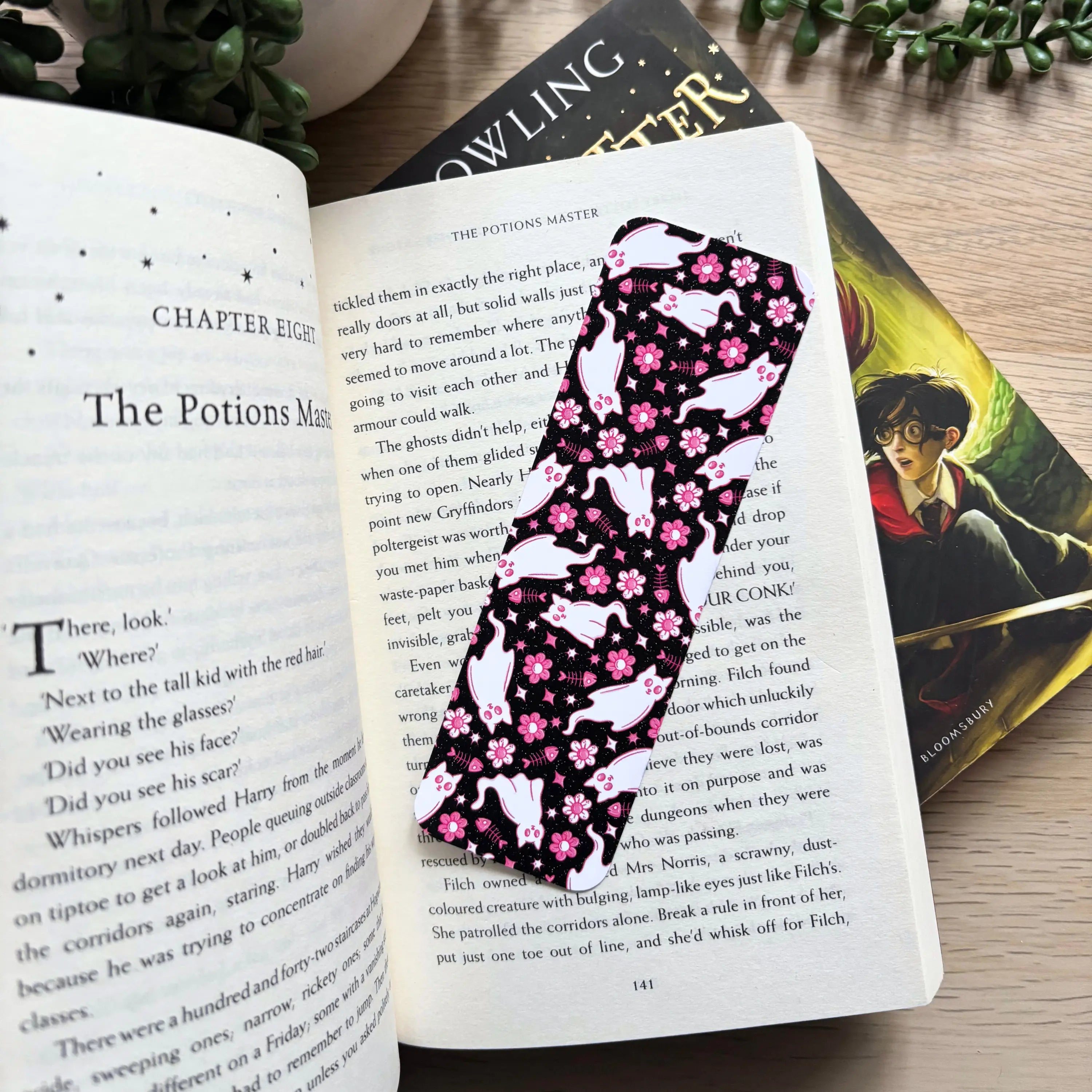 Black metal bookmark with whimsical ghost cat and pink floral design placed inside an open book page.