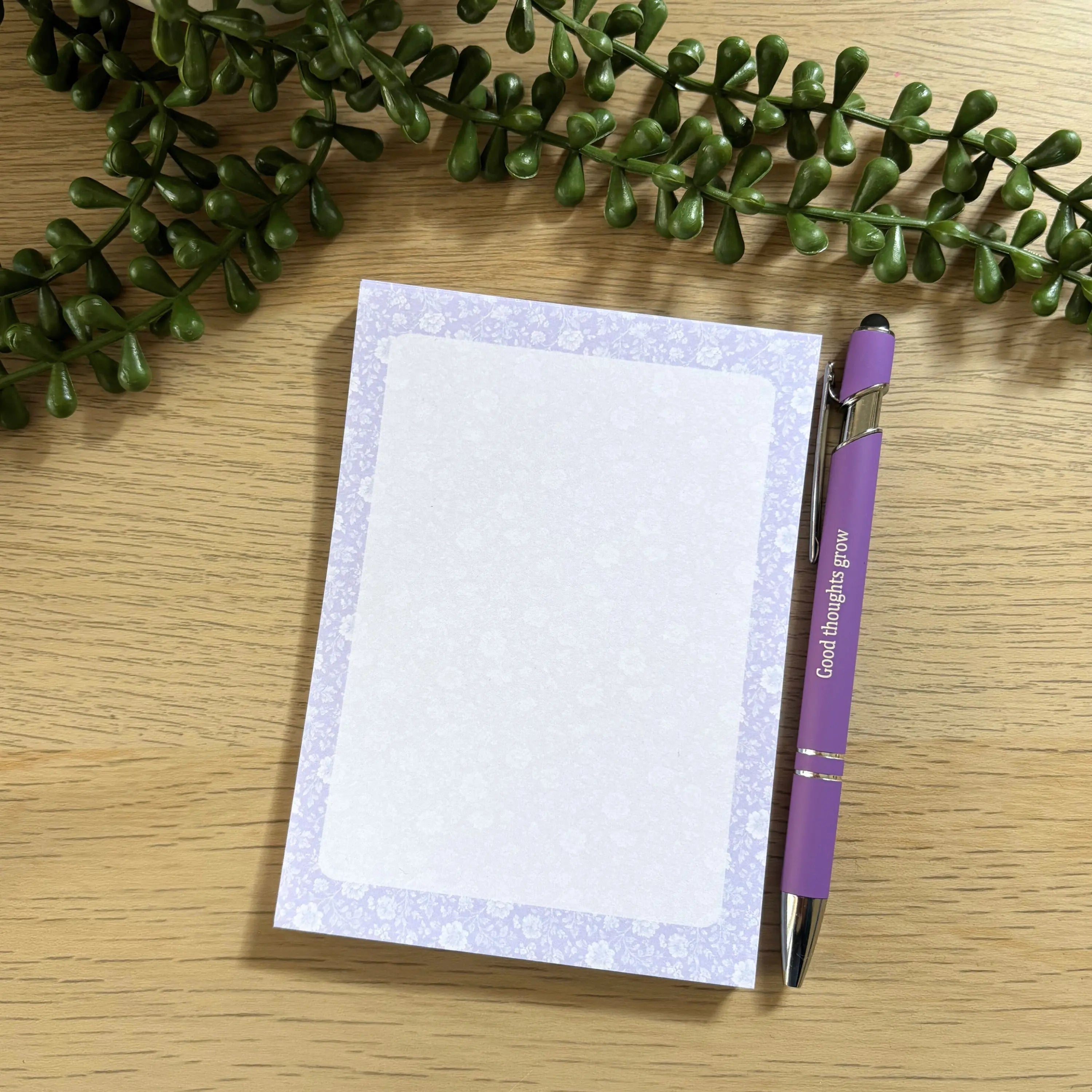 Small lavender floral notepad with white writing space and matching purple pen on wooden surface.