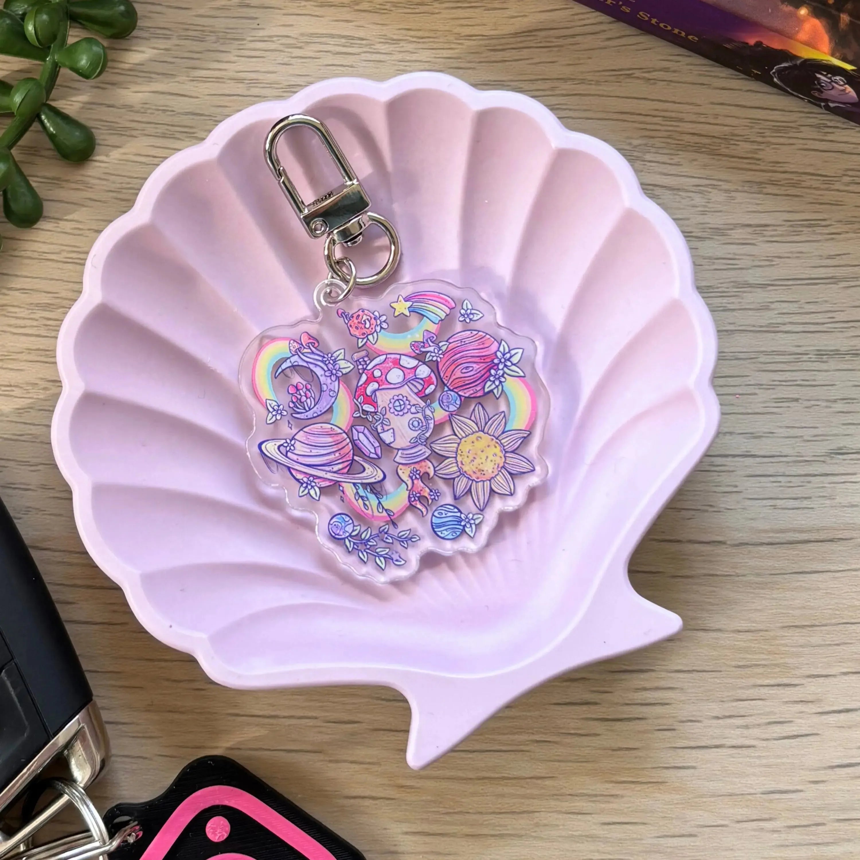 Clear acrylic keyring with colorful whimsical design of planets, flowers, and mushrooms on silver clasp in pink shell dish.