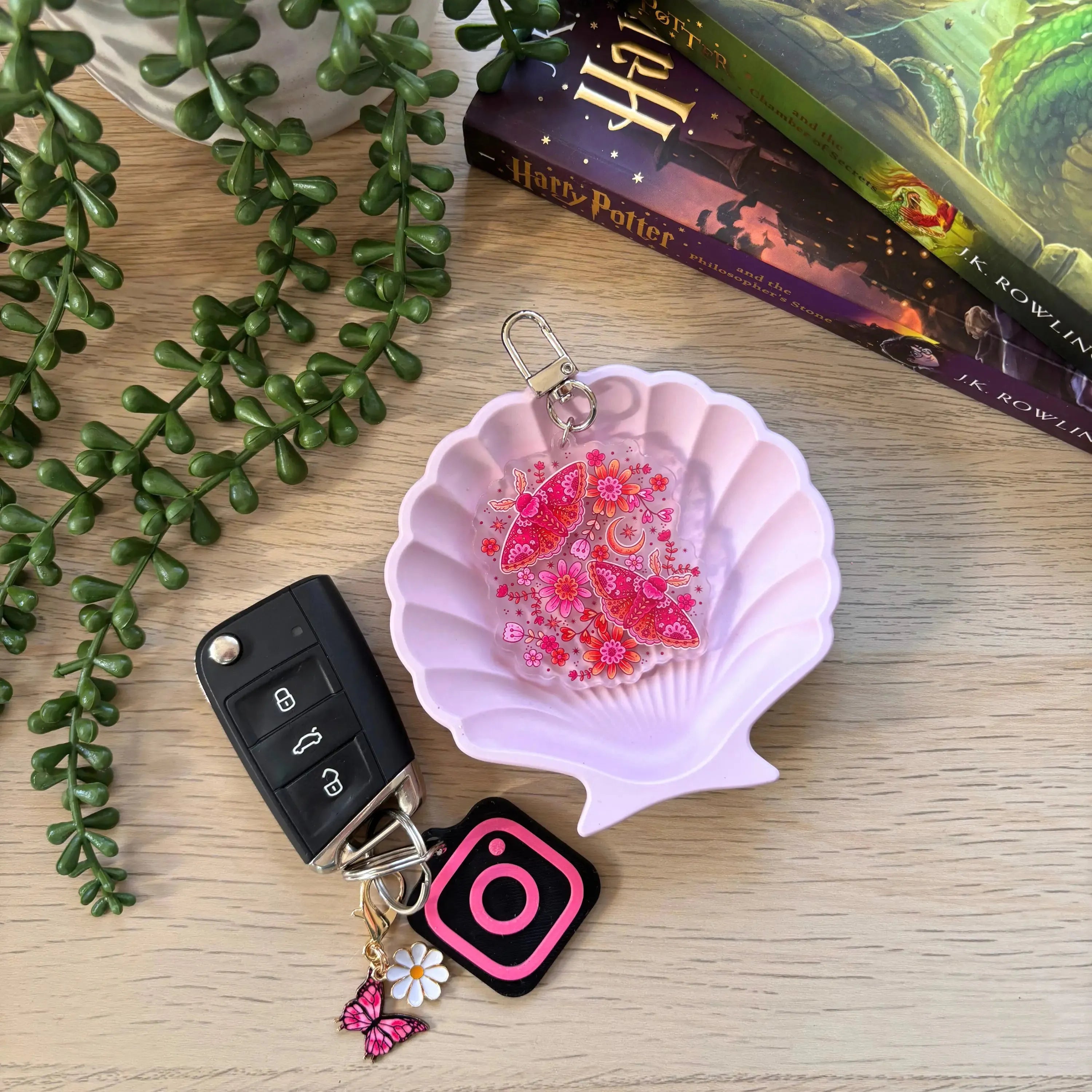 Pink translucent moth keyring with floral details on a light pink seashell dish next to car keys and books.