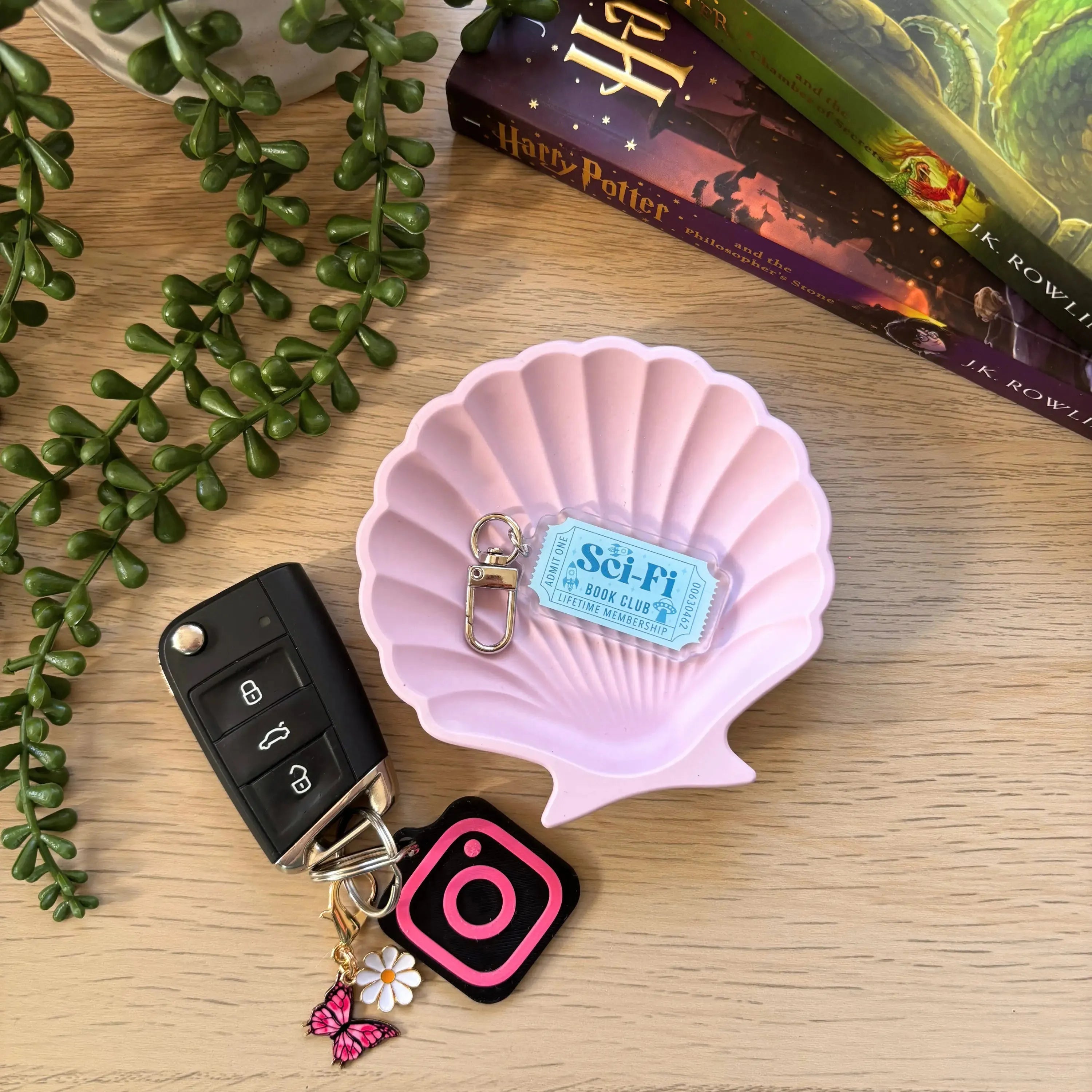 Light blue sci-fi bookclub keyring with clasp attached to black car key and pink Instagram charm on wooden surface next to.