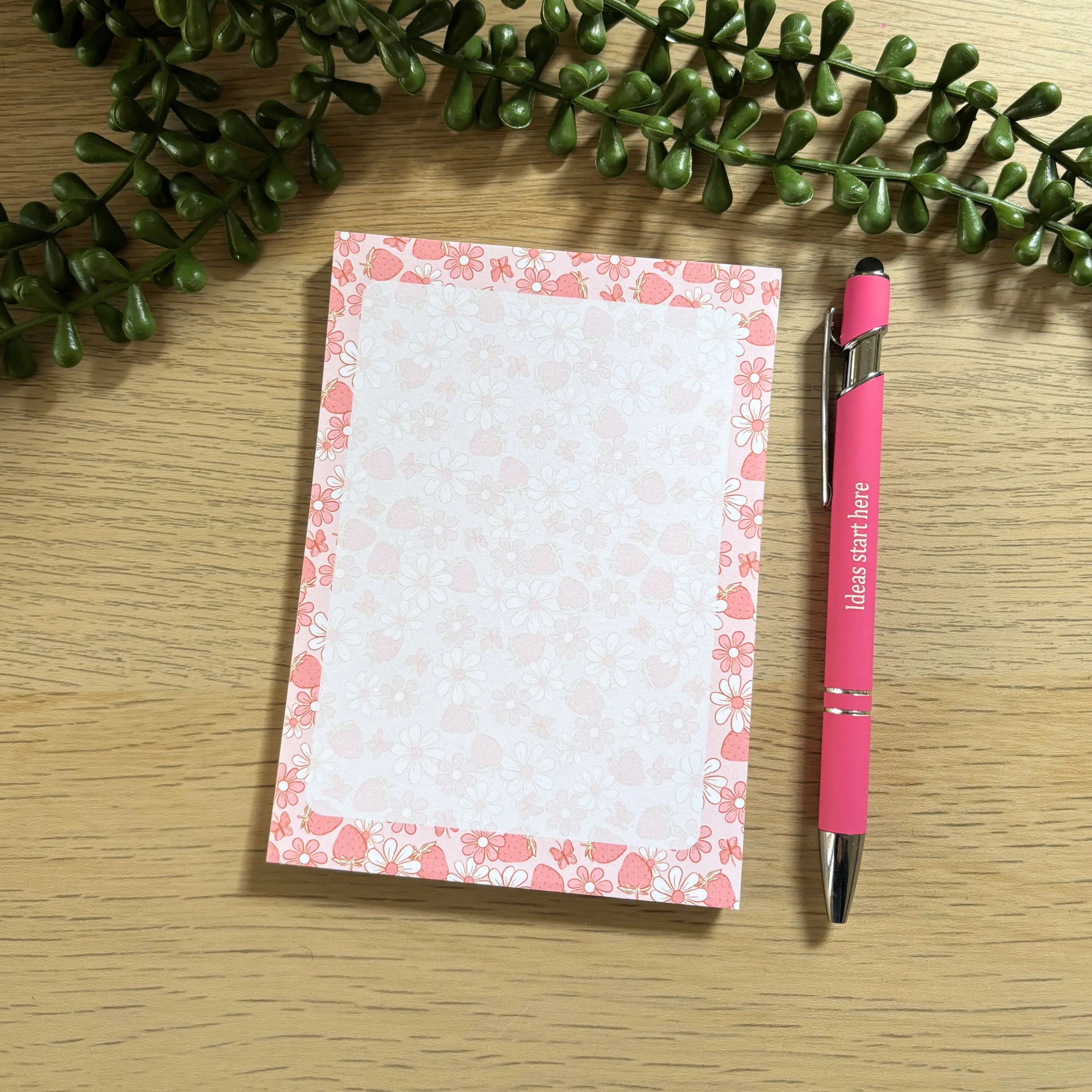 Small Strawberry Field notepad with pink floral border next to pink and black ballpoint pen on wood surface.
