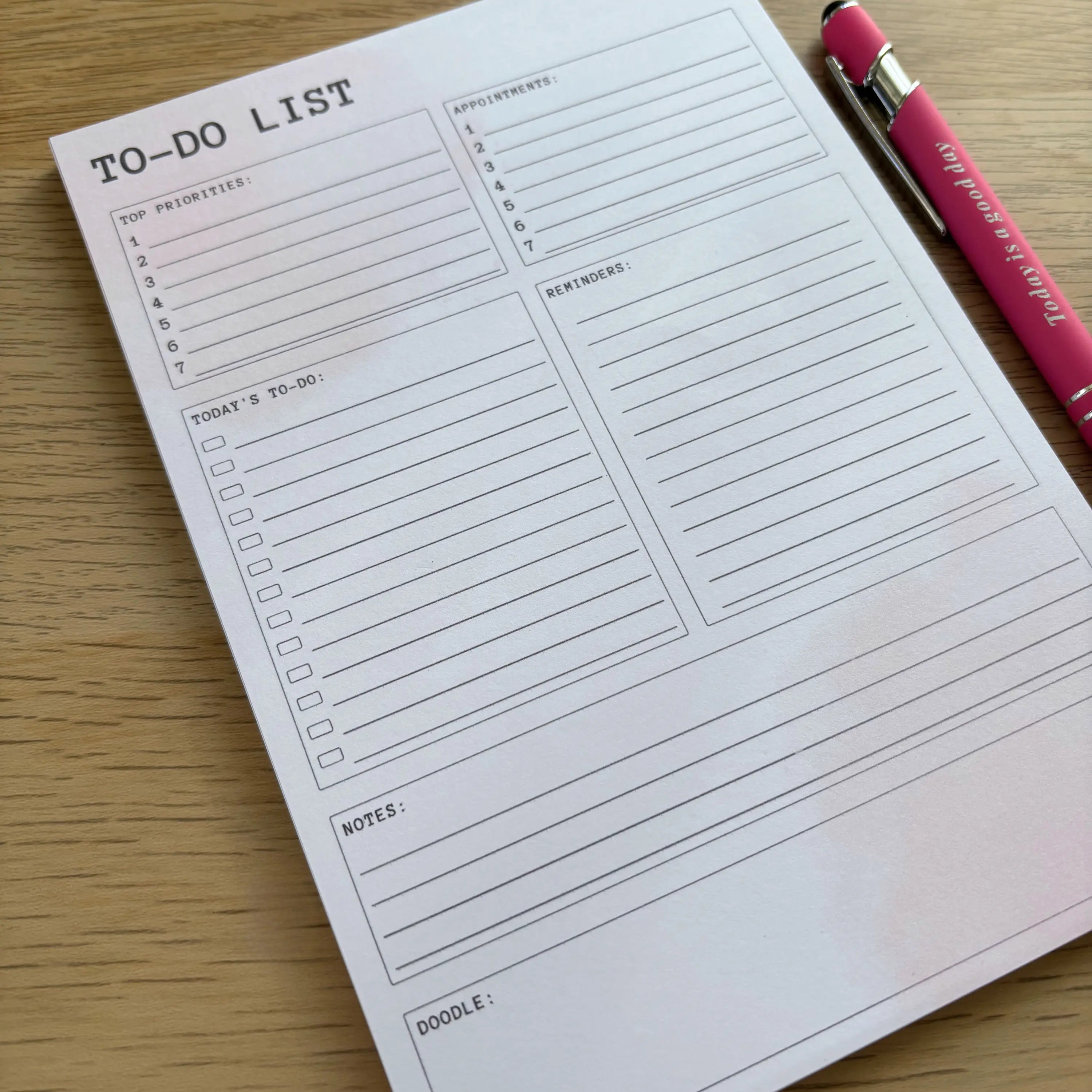 Soft pink to-do notepad with sections for tasks, shopping, priorities, notes, and schedule on wooden surface.