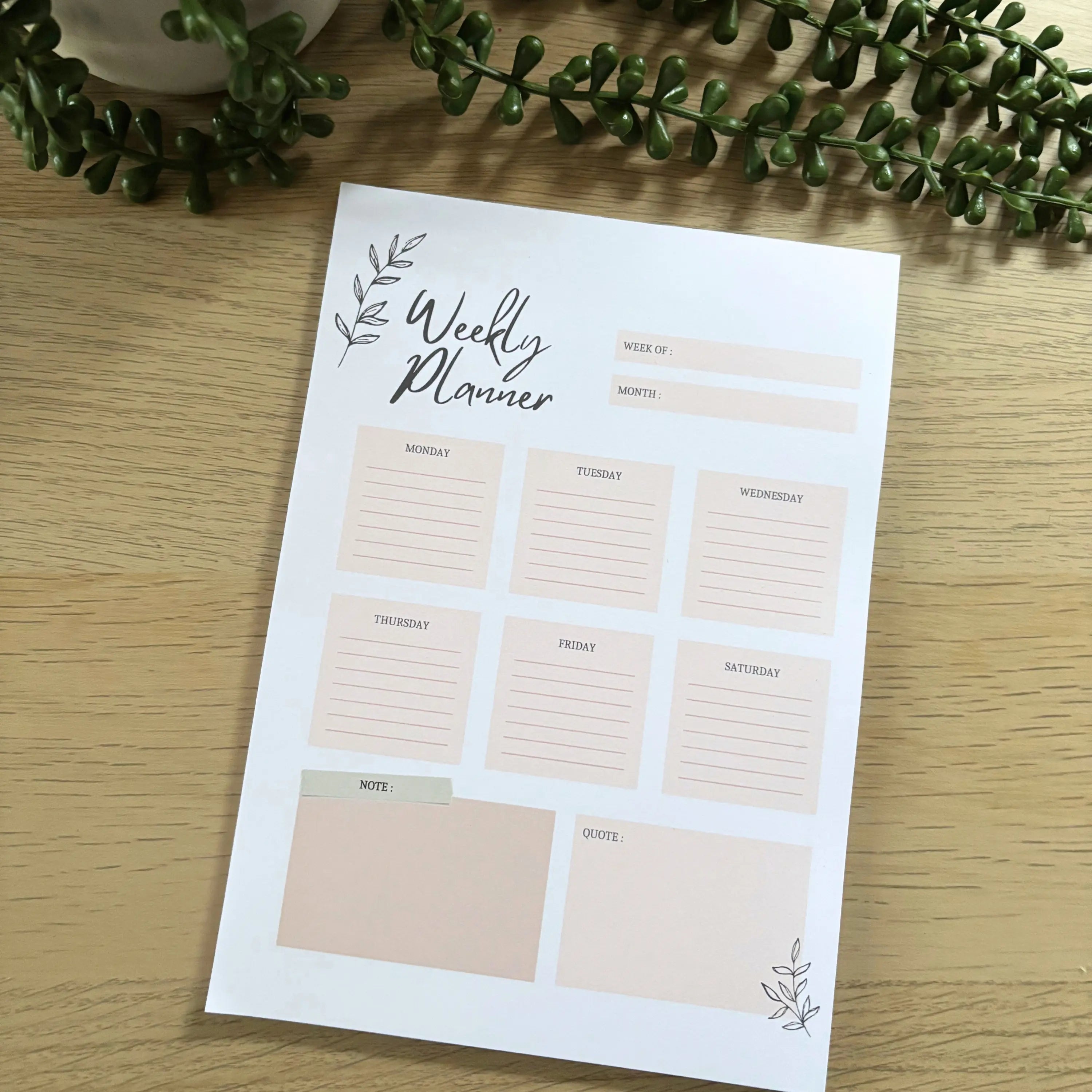 Minimalist weekly planner notepad with seven day blocks and two notes sections on wooden surface with greenery.